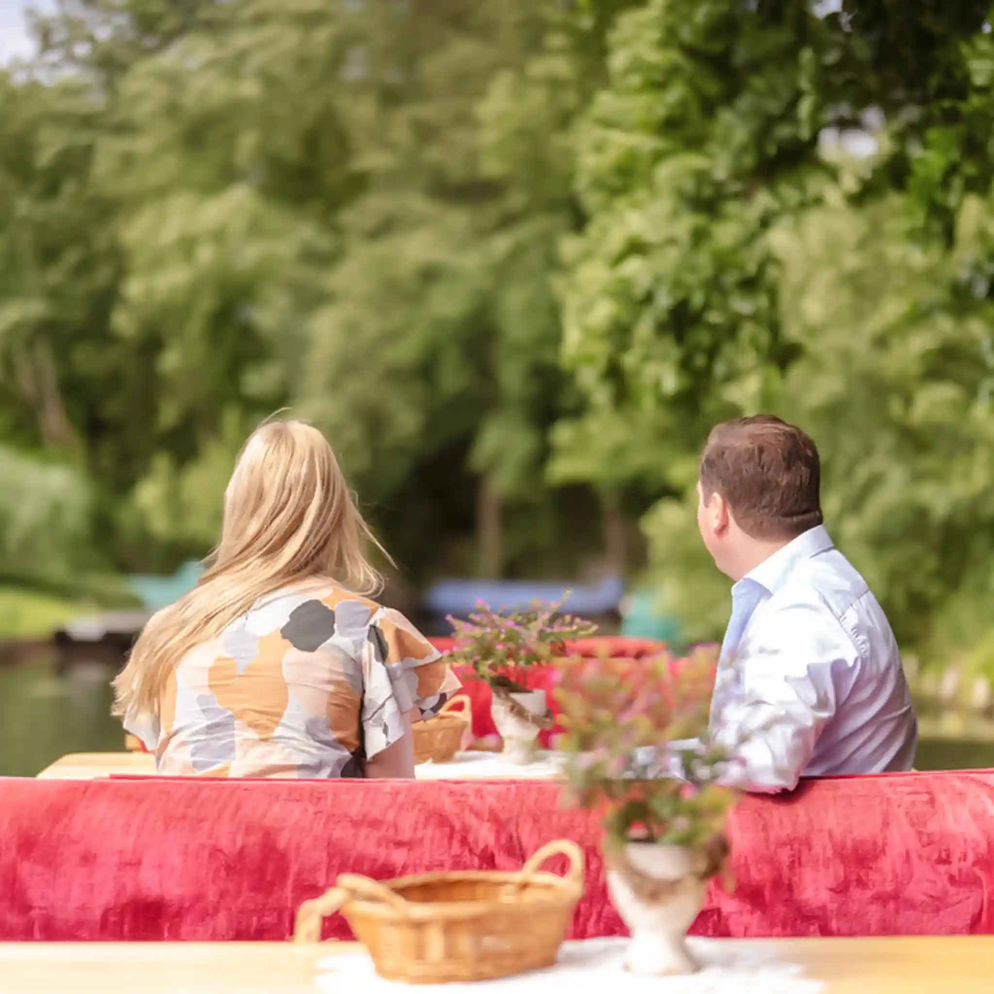 Ein Mann und eine blonde Frau sitzen von hinten gesehen auf einem Kahn mit roten Polstern und fahren durch eine grüne Flusslandschaft.