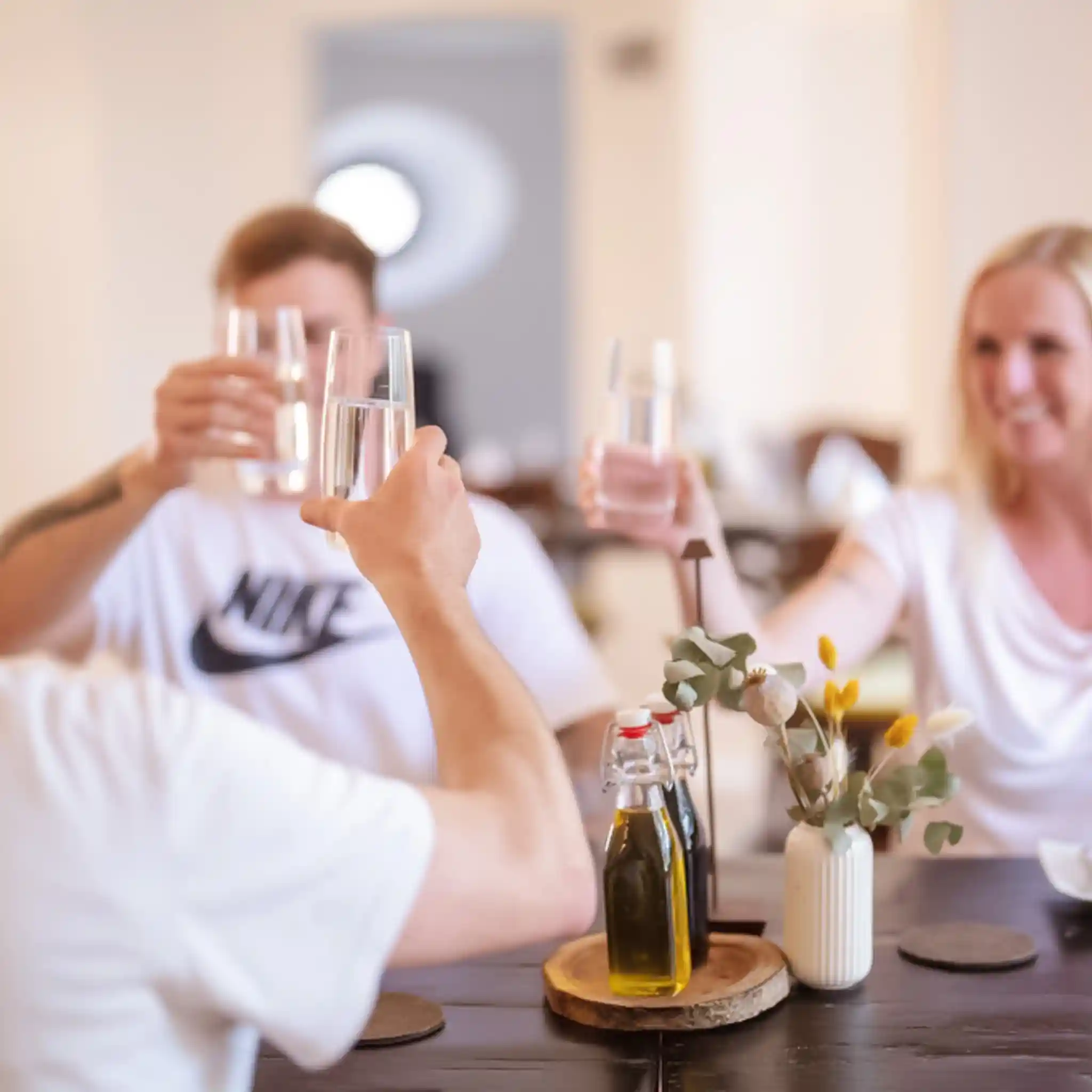 Drei Personen sitzen an einem Tisch und stoßen gut gelaunt mit Wassergläsern an.