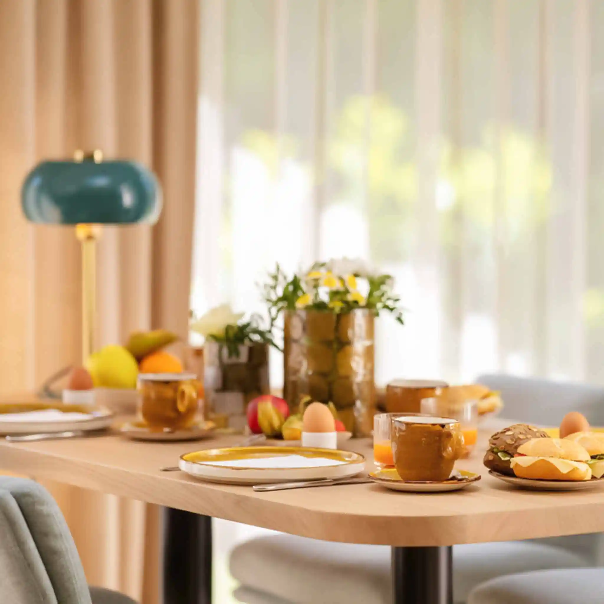 Ein reichlich gedeckter Frühstückstisch mit Brötchen, gekochten Eiern, Tassen und frischen Blumen vor einem hellen Fenster.