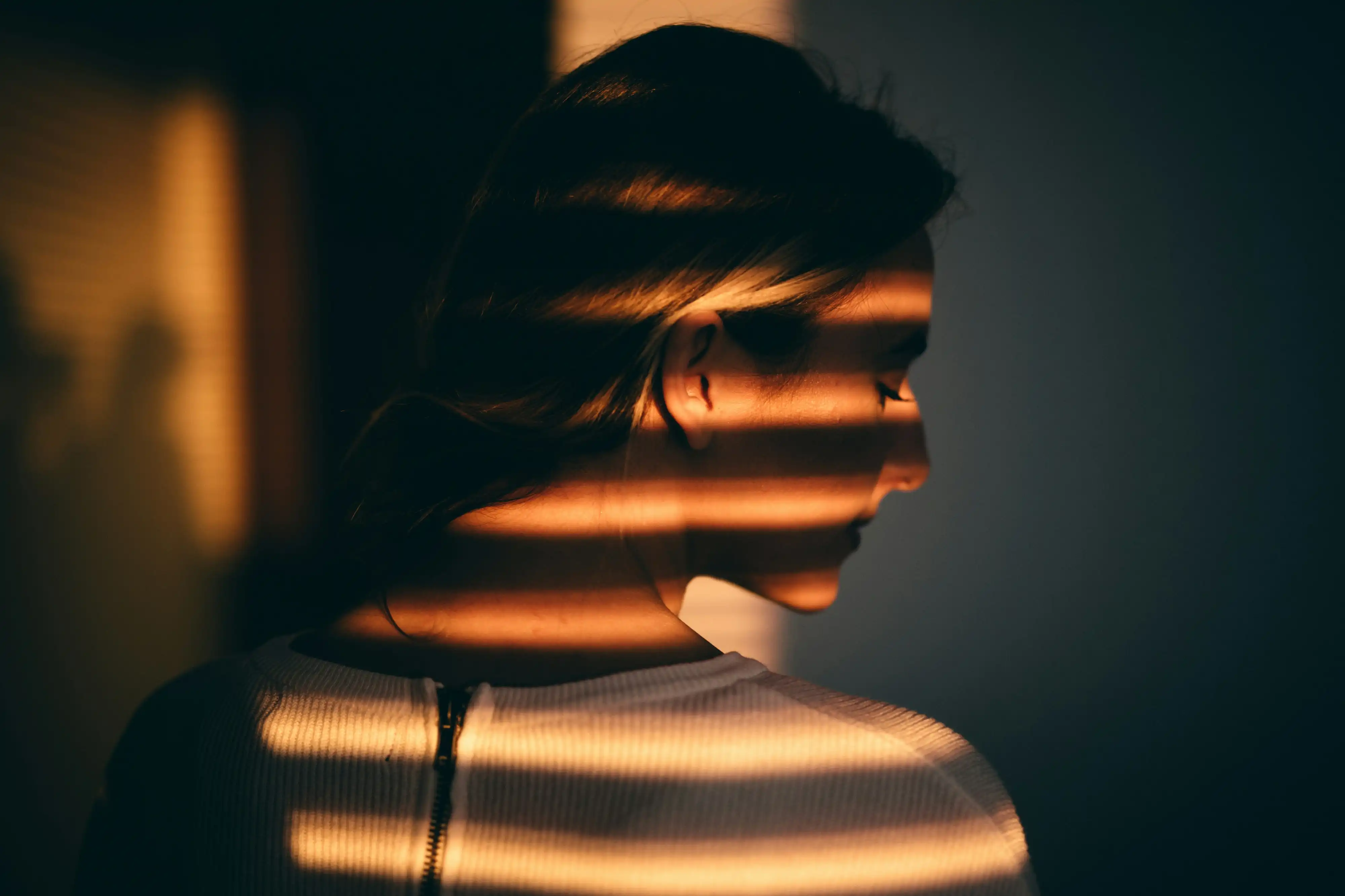 Profilansicht einer Frau von hinten im weißen Oberteil, auf die warmes Sonnenlicht durch eine Jalousie gestreift fällt.