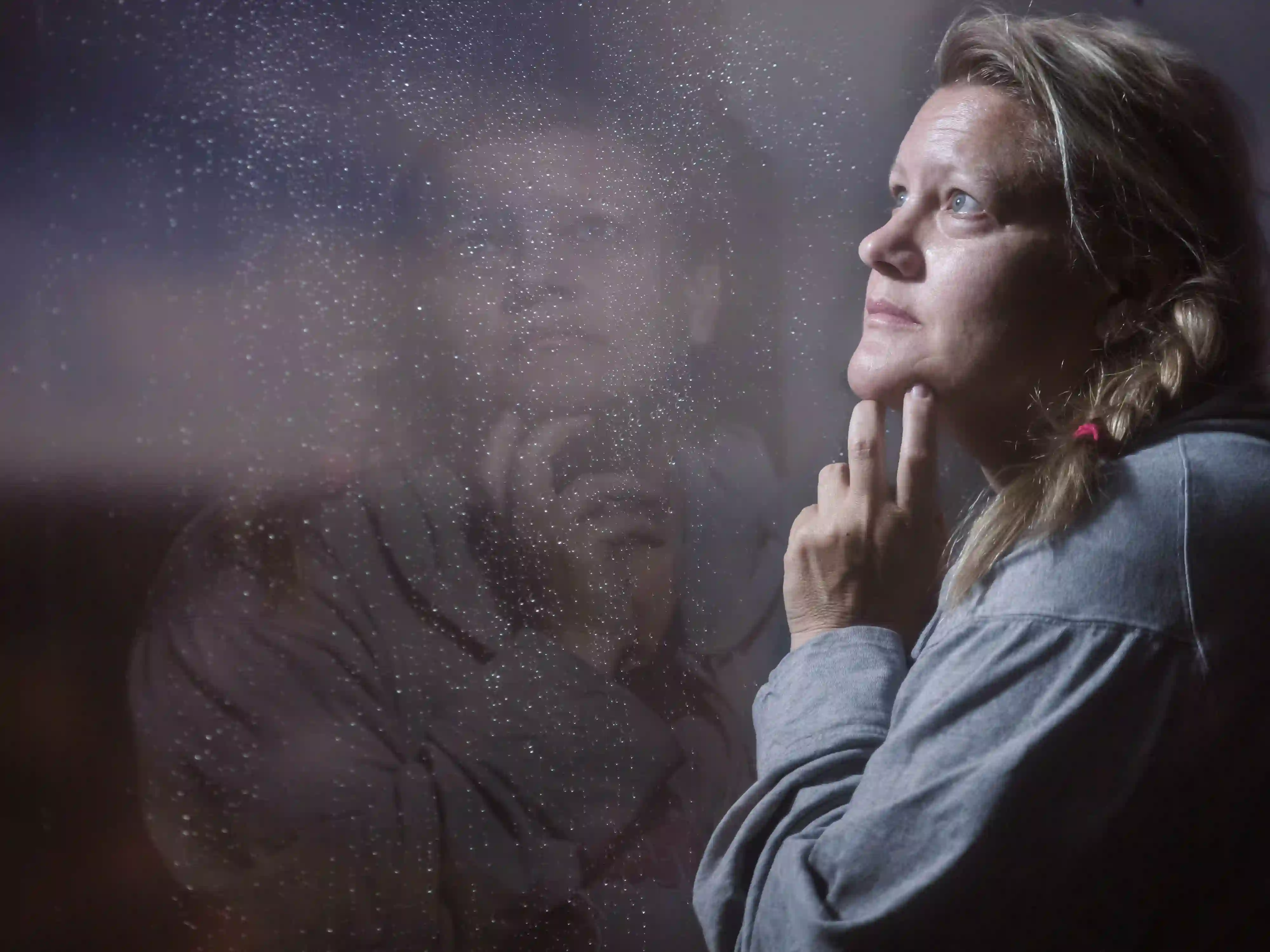 Eine blonde Frau blickt nachdenklich durch eine regennasse Scheibe, in der sich ihr Bild spiegelt.