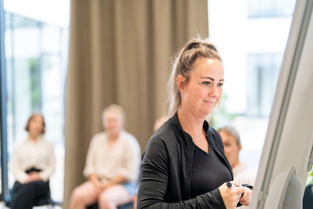 Eine lächelnde Frau mit Zopf im schwarzen Sportshirt steht an einem Flipchart vor einer Zuhörergruppe.