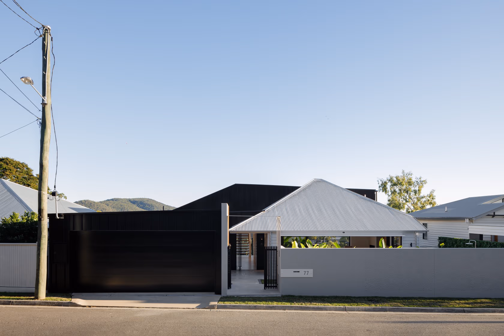 A minimalist house exterior showcases a black garage door, a light-colored roof, and a blend of white and gray walls, surrounded by greenery and hills in the background..
