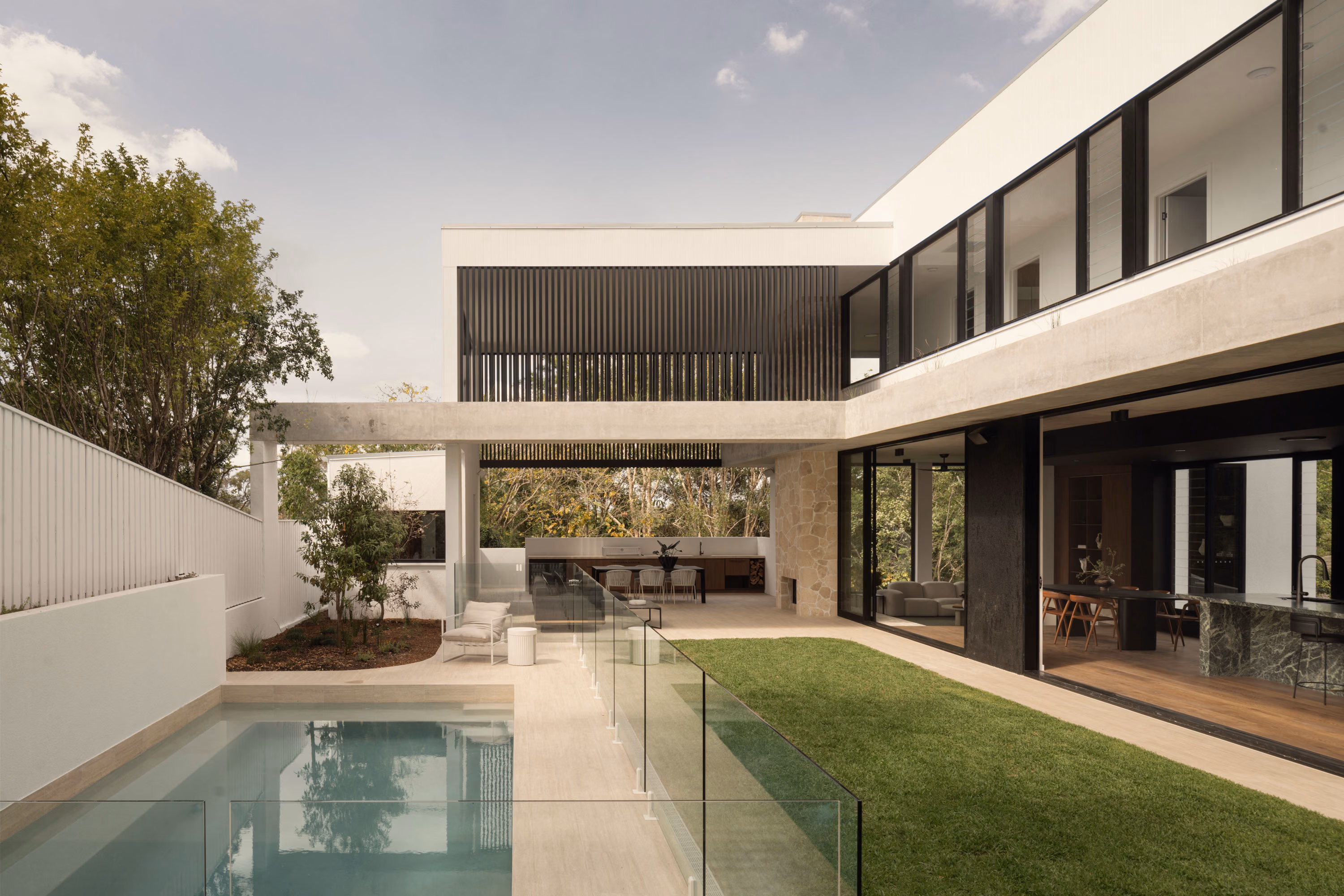 Rear courtyard view of Stanley featuring a lap pool, glass pool fence, landscaped lawn, and open-plan living spaces framed by concrete beams and black vertical screening.