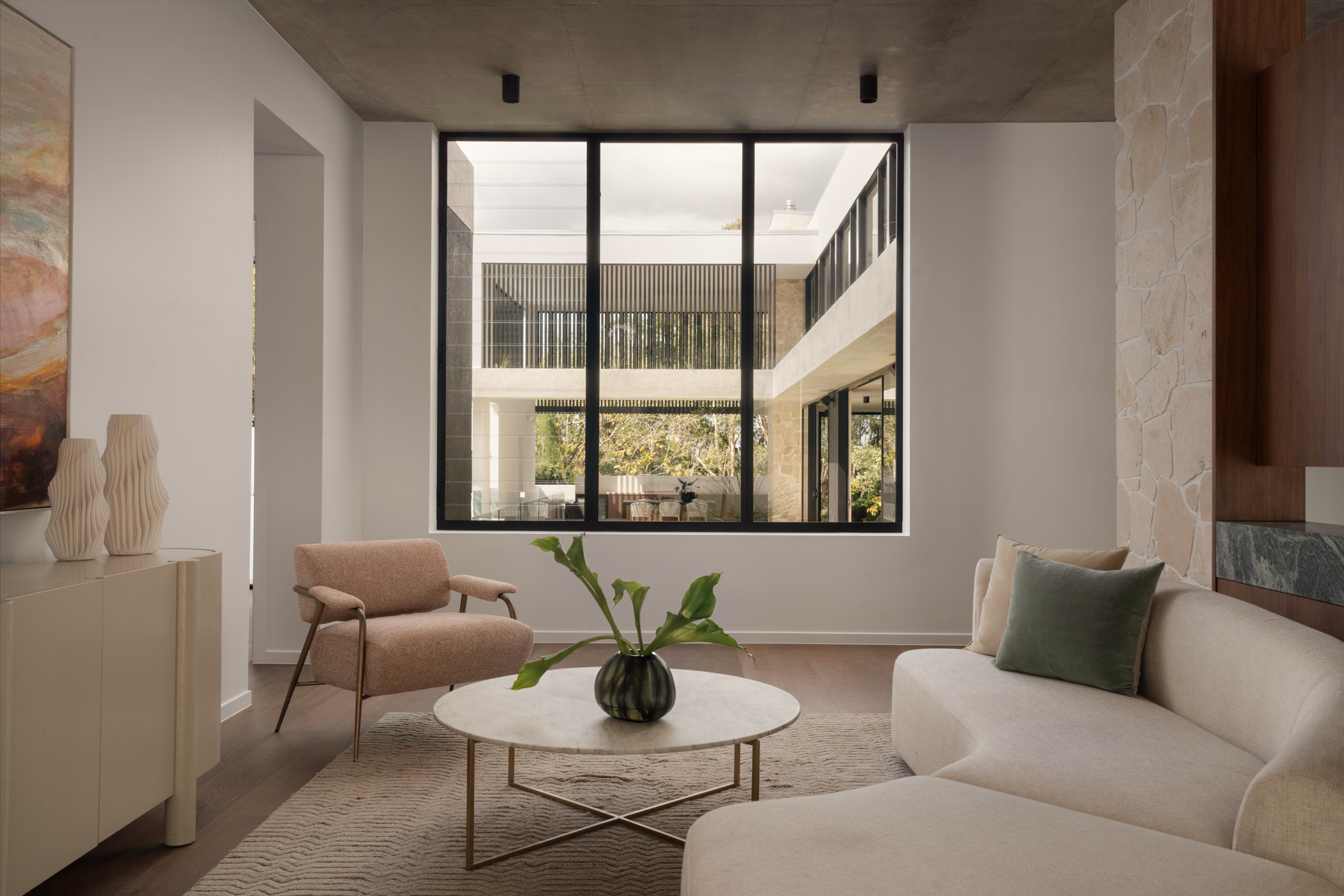 Cozy sitting area in Stanley with a curved sofa, textured armchair, marble coffee table, and large picture window overlooking the central courtyard.