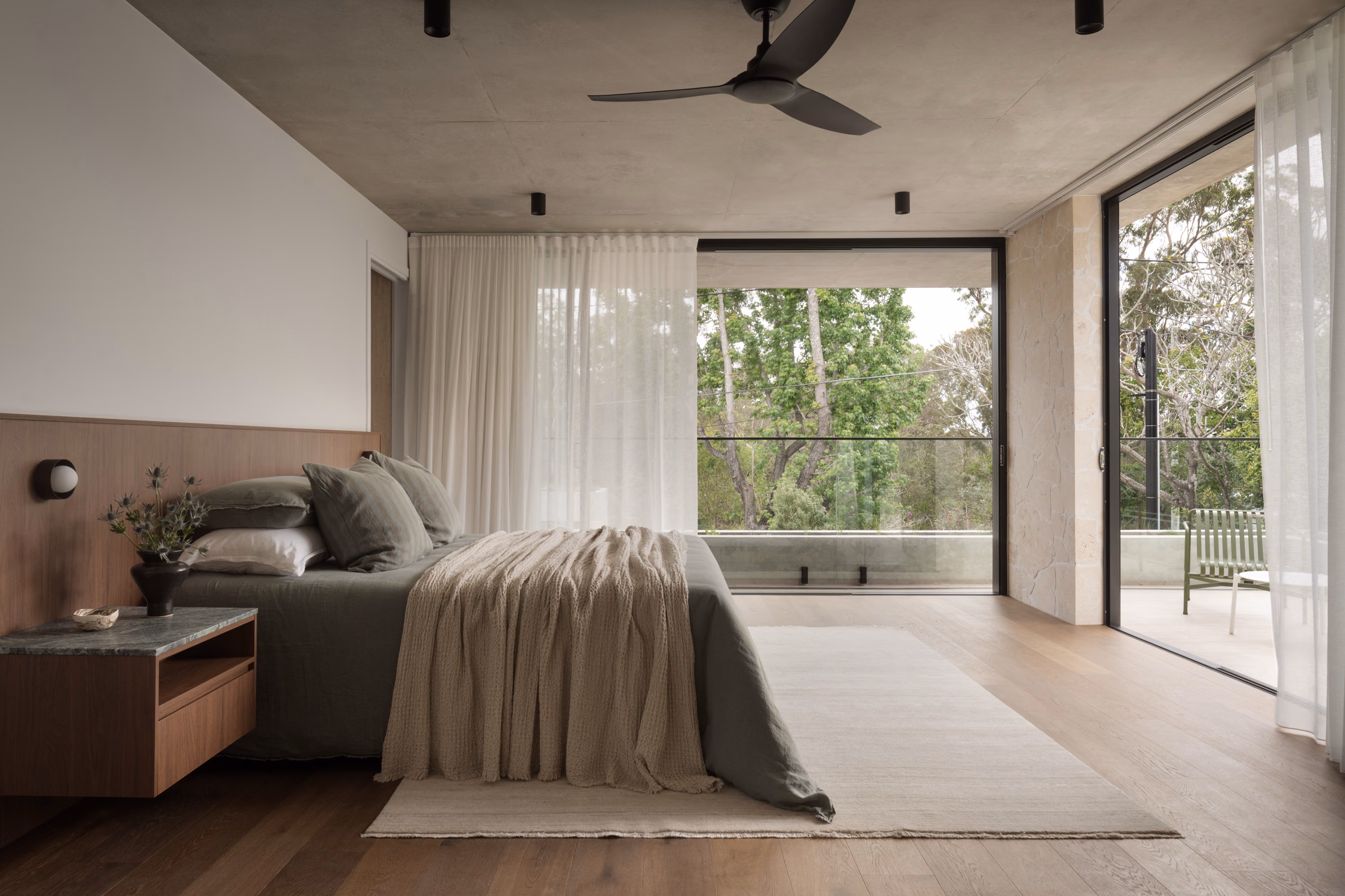 Primary bedroom at Stanley with a walnut bedhead, soft linen bedding, floor-to-ceiling glazing, and access to a private balcony surrounded by trees.