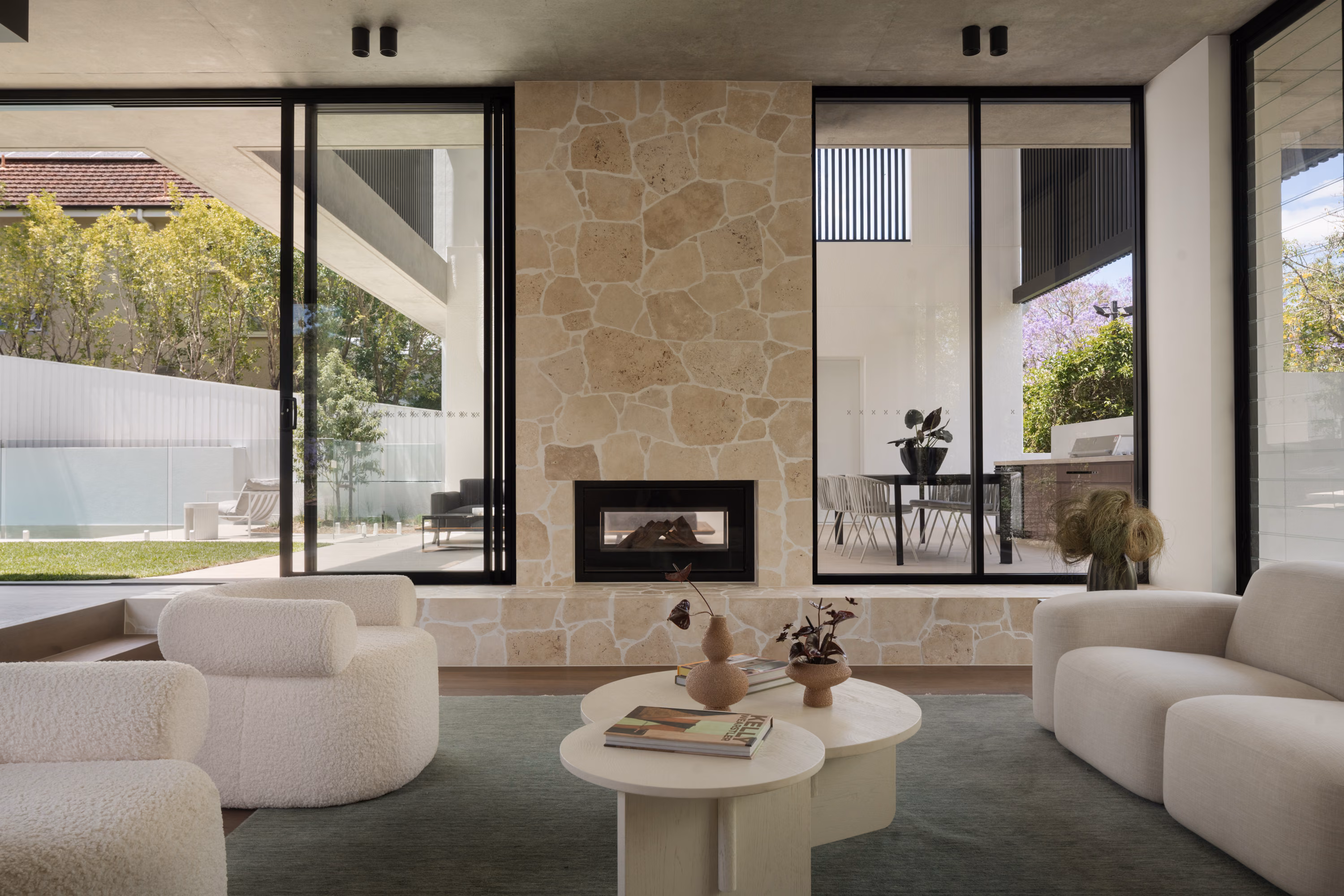 Stone fireplace in Stanley’s living room framed by full-height glazing with views to the courtyard, outdoor dining area, and pool.
