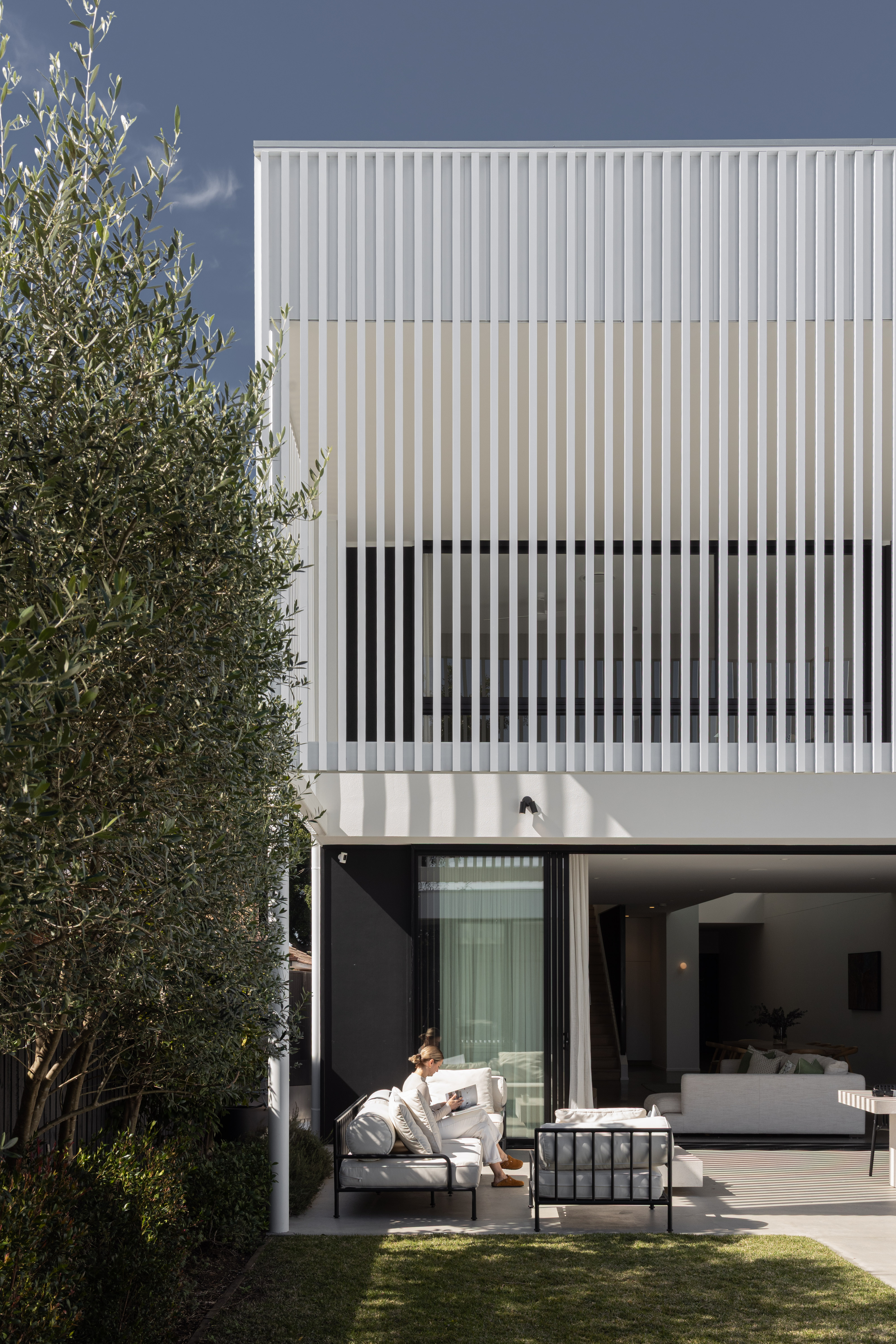 Rear elevation at Burilda featuring the white louvre screen upper level, open alfresco with outdoor lounge, glass pool fence, and person reading on sofa