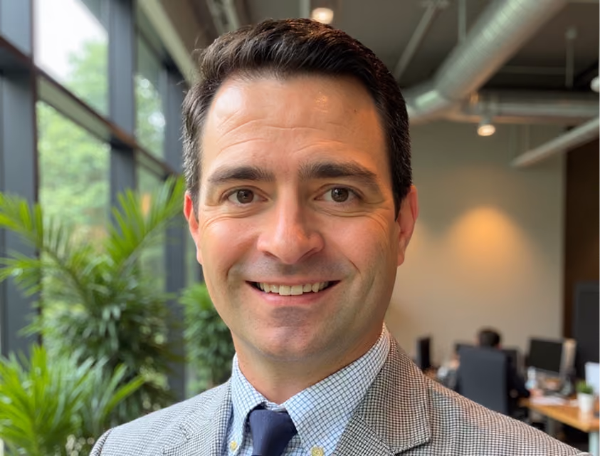 Smiling man with dark hair wearing a light gray checkered suit jacket, white shirt, and dark tie in a modern office with large windows and plants.
