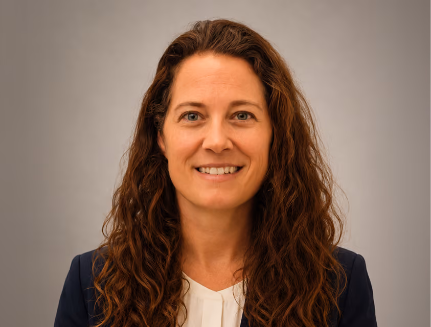 Smiling woman with long curly brown hair wearing a white blouse and navy blazer against a neutral gray background.