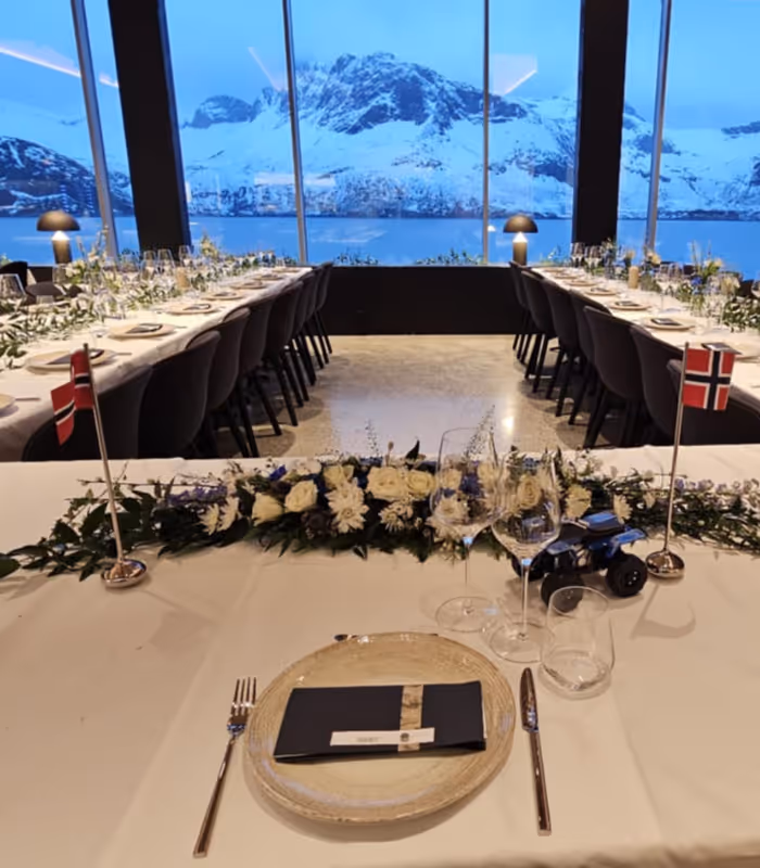Elegantly set dining tables with white floral arrangements and Norwegian flags in a room overlooking snow-covered mountains and a fjord through large windows.