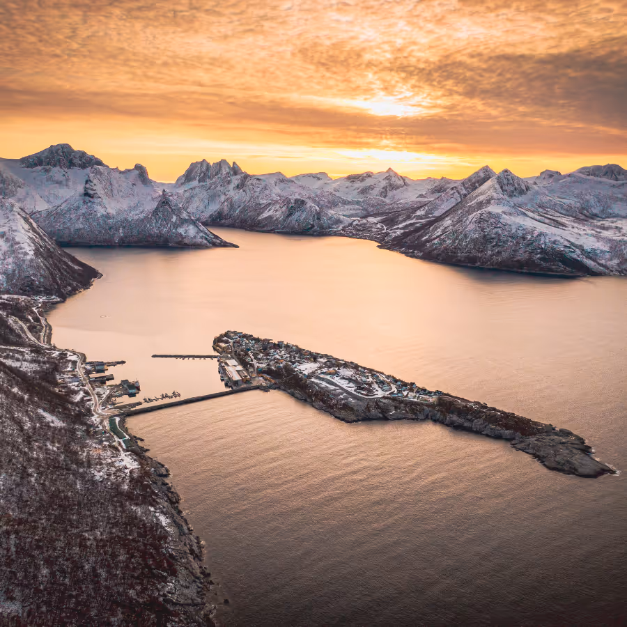 Aerial view of a snowy coastal village on a peninsula surrounded by calm water and rugged snow-covered mountains during a golden sunset.