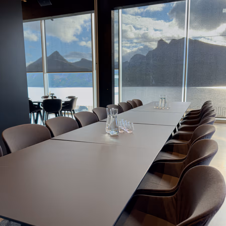 Modern conference room with a long table, chairs, water pitchers, and a large window revealing a mountain and lake view.