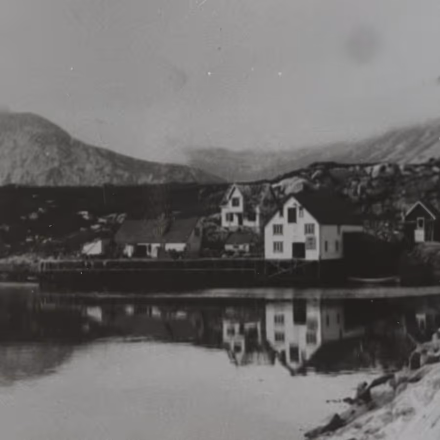 Black and white photo of small houses by a calm lake reflecting the buildings and surrounding rocky hills.