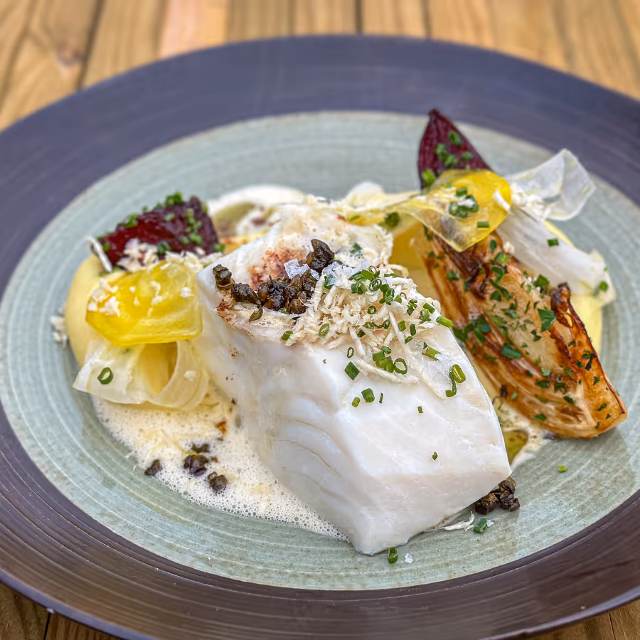 Plate with a piece of cooked white fish garnished with chopped herbs, foam sauce, roasted vegetables, and thin vegetable slices.