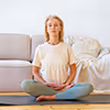 Frau in bequemer Kleidung sitzt mit gekreuzten Beinen meditierend auf dem Boden vor einem Sofa.
