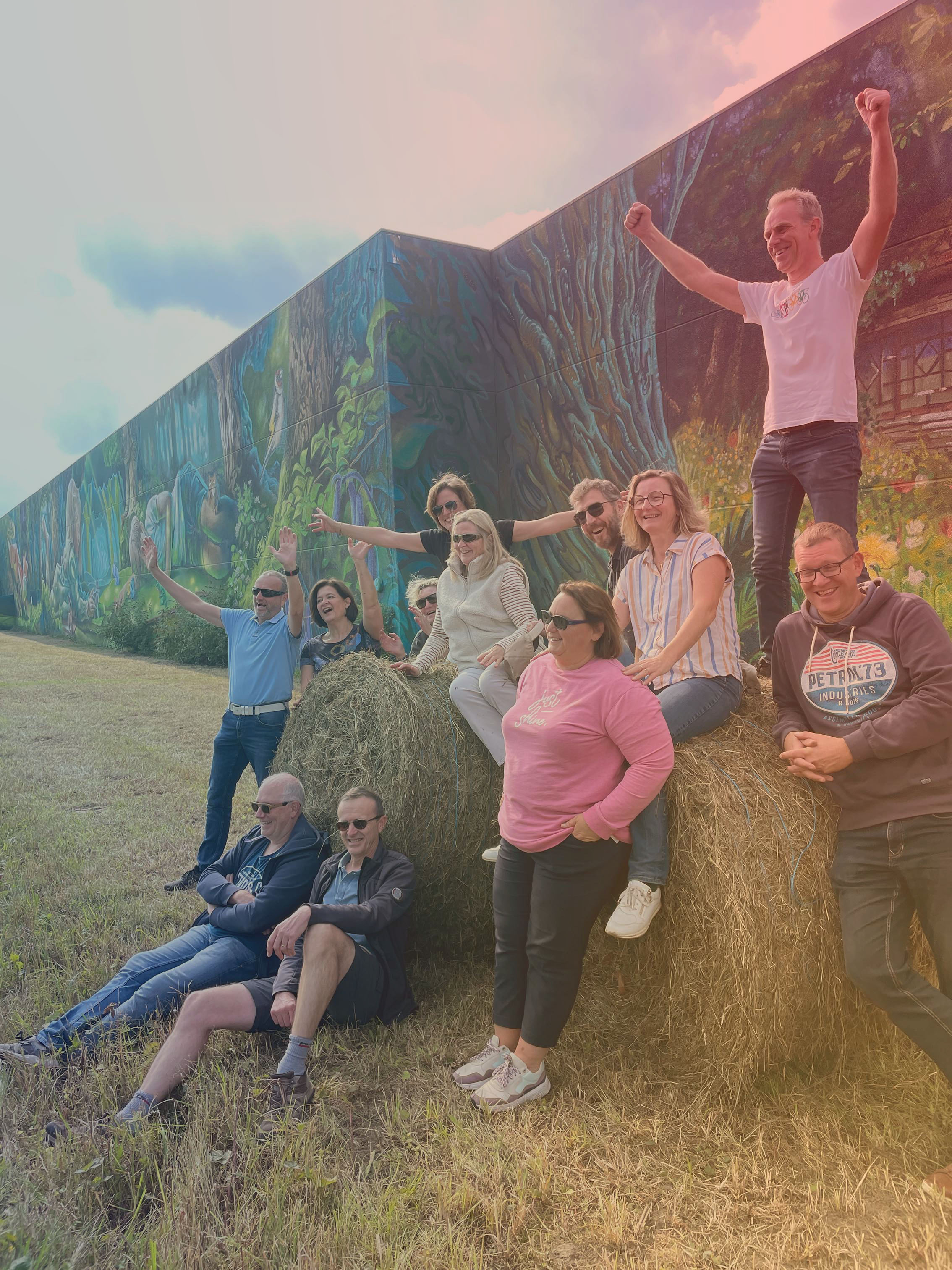 Groep mensen lacht en viert buiten op grote hooirollen met een kleurrijke graffiti-muurschildering op de achtergrond.