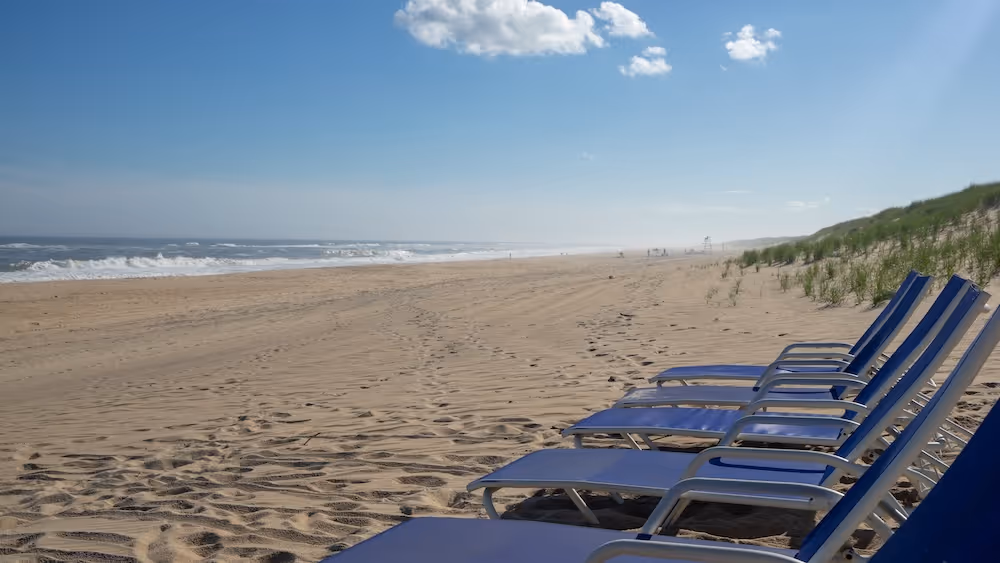 Row of blue beach chairs on sandy shore with ocean waves and a clear sky with a few clouds.