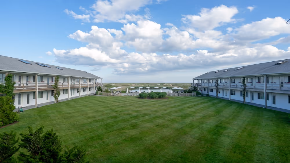 Two white two-story buildings with balconies facing a large green lawn under a partly cloudy blue sky.