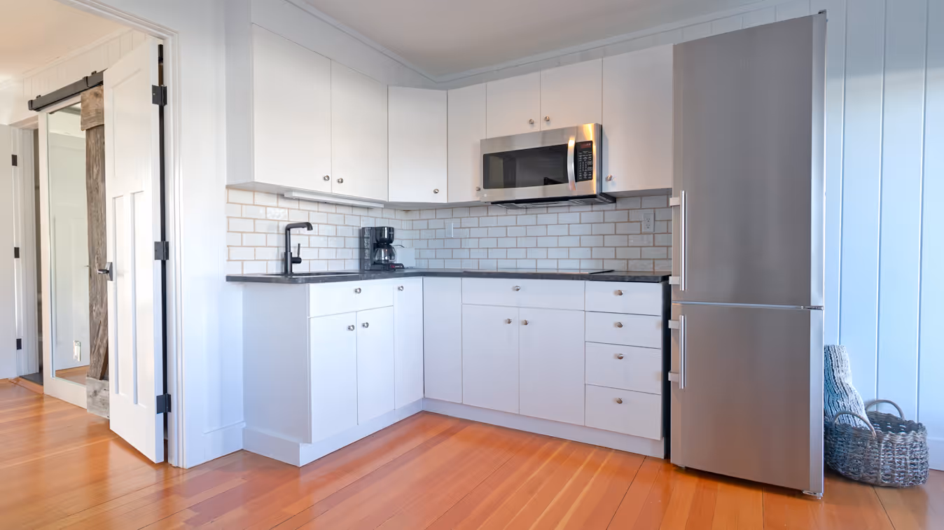 Modern white kitchen corner with black countertop, stainless steel microwave and refrigerator, wooden floor, and white subway tile backsplash.
