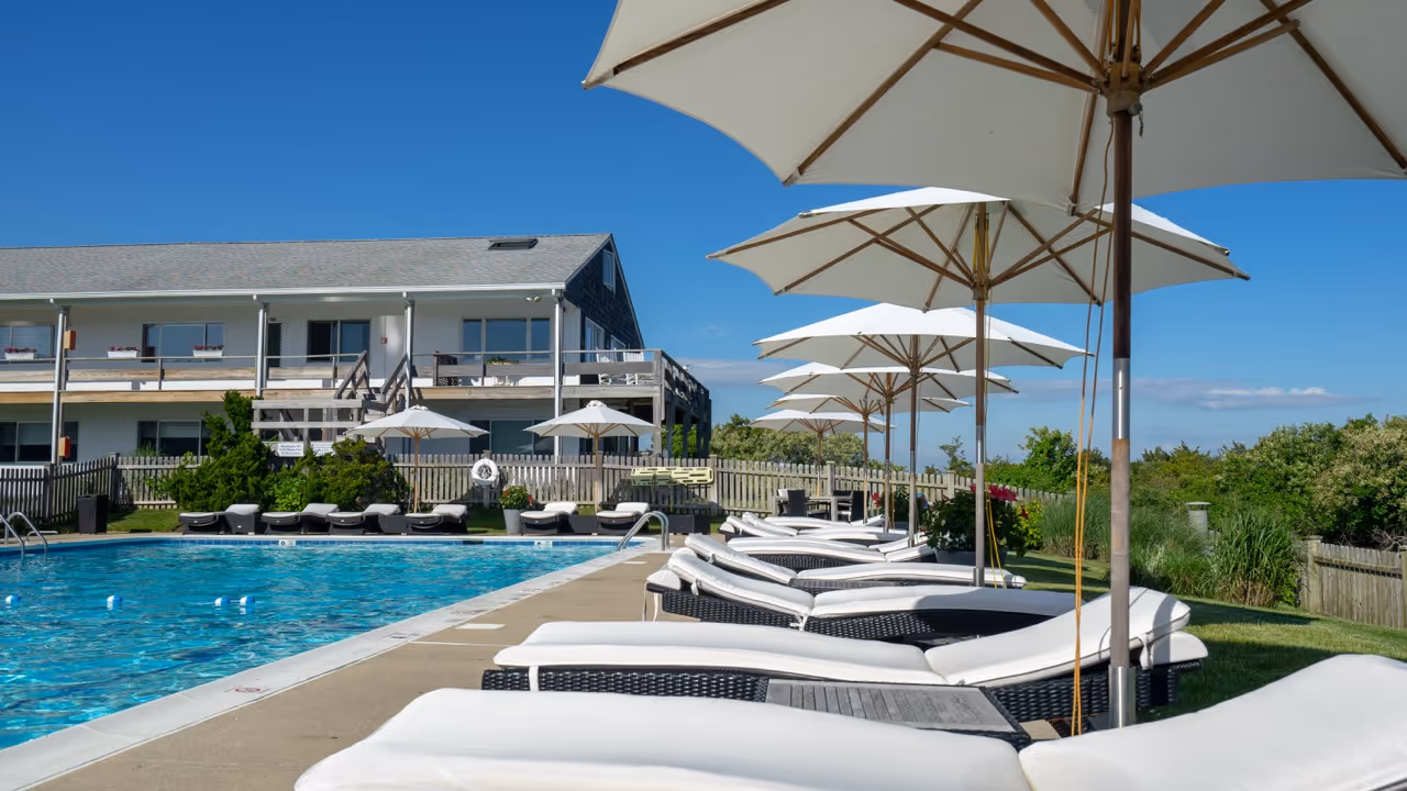 Outdoor swimming pool with lined white cushioned lounge chairs and large white umbrellas on a sunny day.