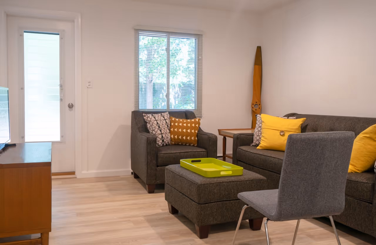 Modern living room with gray armchair, matching ottoman with green tray, gray sofa with yellow pillows, wooden side table, and a wooden propeller decoration by the window.