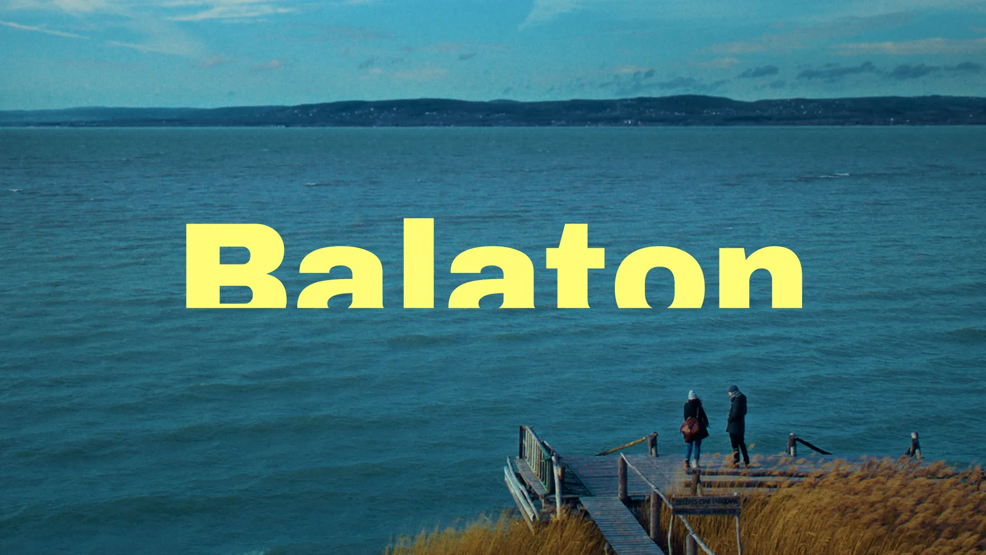Two people standing on a wooden pier overlooking a large blue lake with distant hills under a cloudy sky.