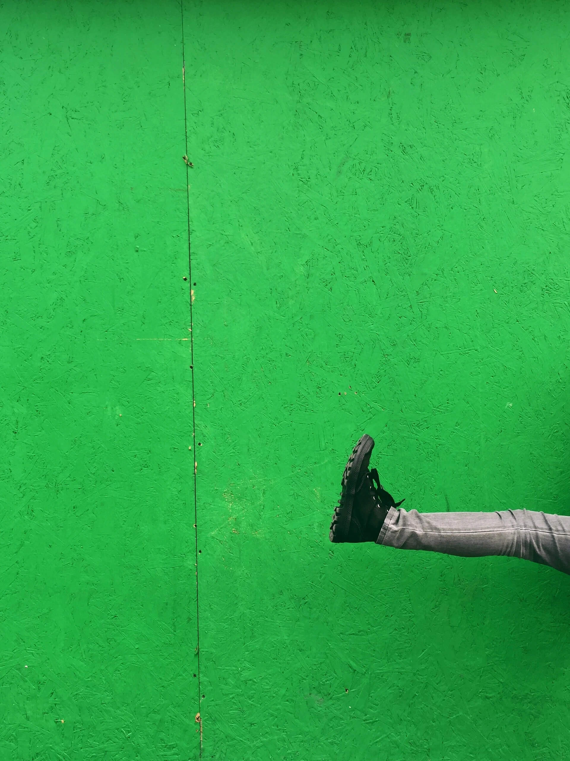 A man in black jeans and a black shirt leaps against a vibrant green wall, showcasing dynamic movement and energy.
