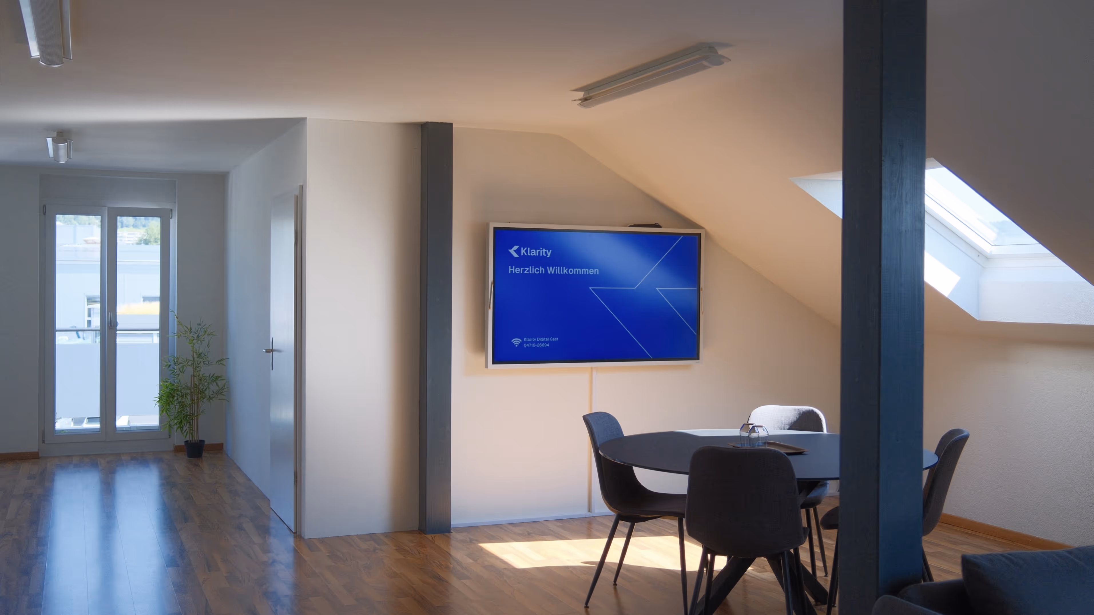 Büro von Klarity Digital Moderner Besprechungsraum mit rundem Tisch, vier Stühlen, einer grünen Pflanze, Dachfenster und Bildschirm mit Willkommensnachricht.