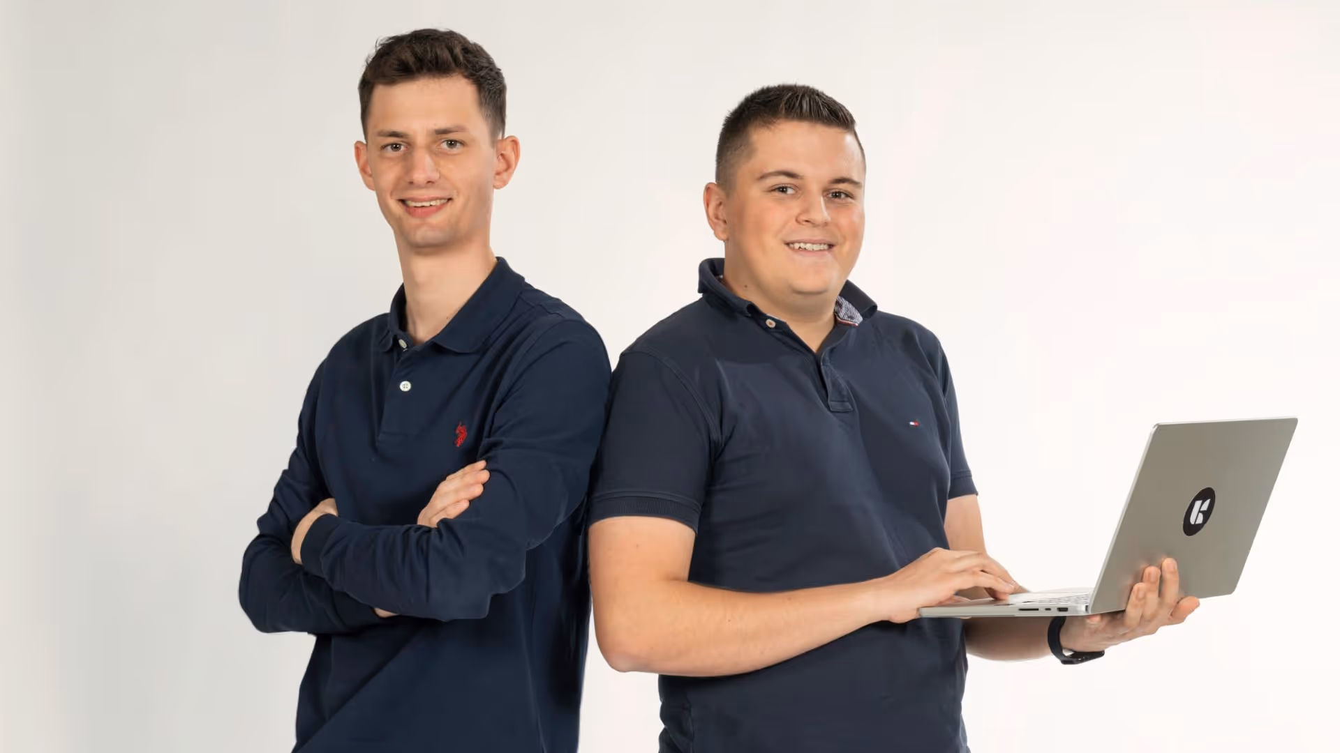 Zwei junge Männer in dunkelblauen Poloshirts lächeln, einer steht mit verschränkten Armen, der andere hält einen Laptop mit einem Logo.