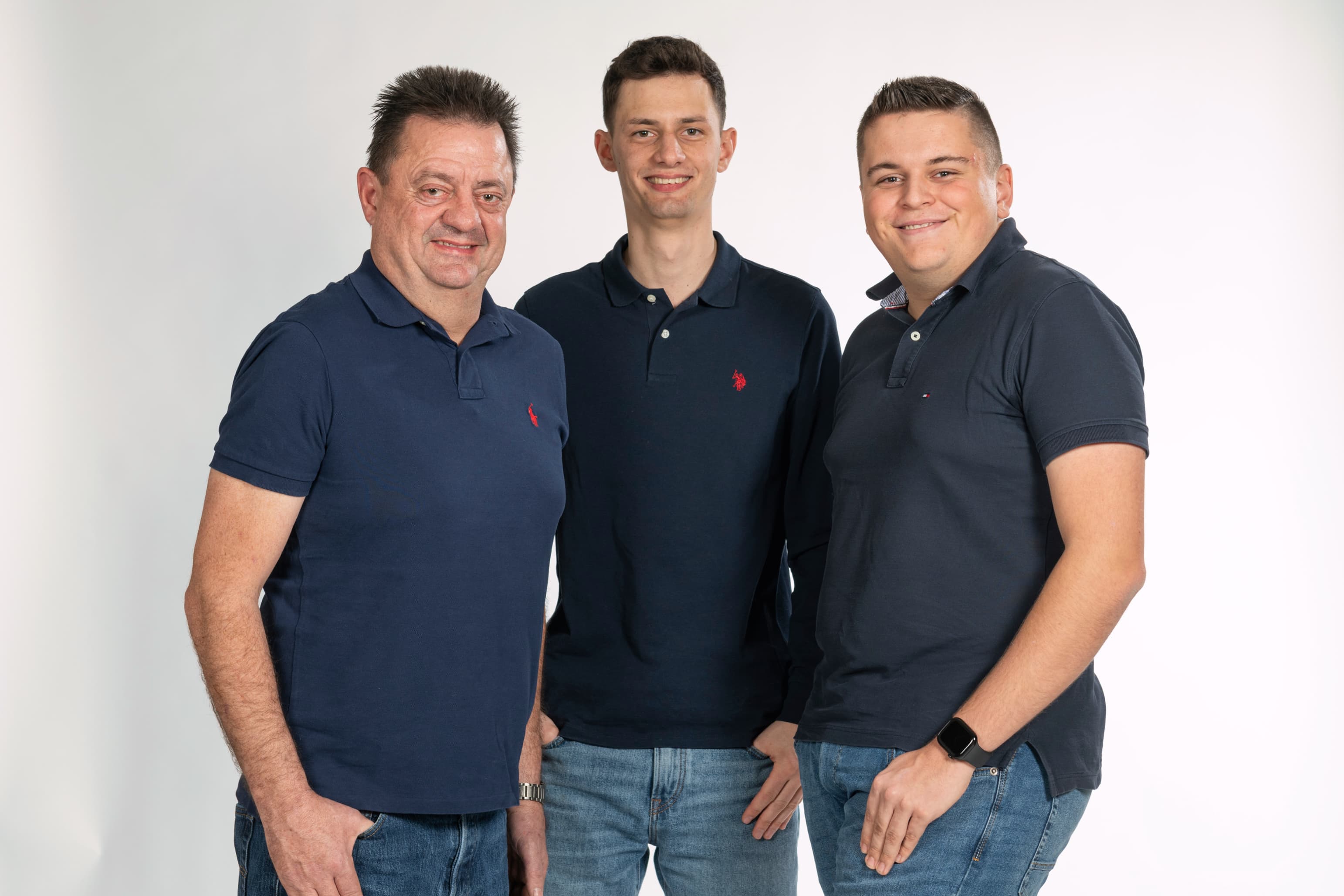 Drei Männer in dunkelblauen Poloshirts und Jeans posieren lächelnd vor weissem Hintergrund.