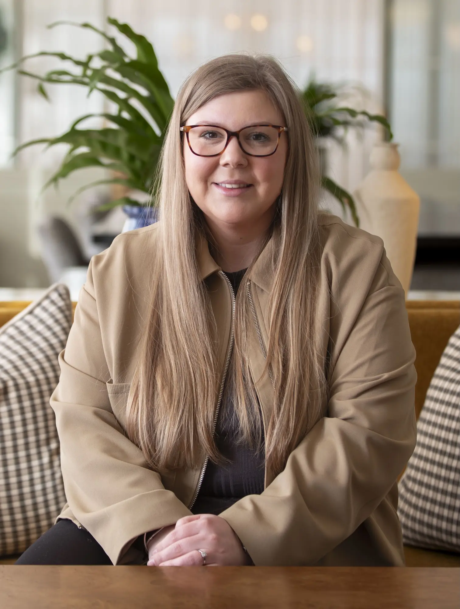 Woman with long blonde hair and glasses wearing a tan jacket, sitting with hands crossed on a wooden table, with green plants and checkered cushions in the background.