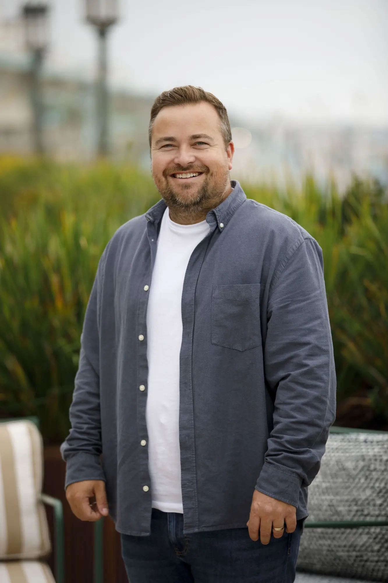 Smiling man with short brown hair and beard wearing a white t-shirt under a dark gray button-up shirt, standing outdoors with green plants and blurred background.