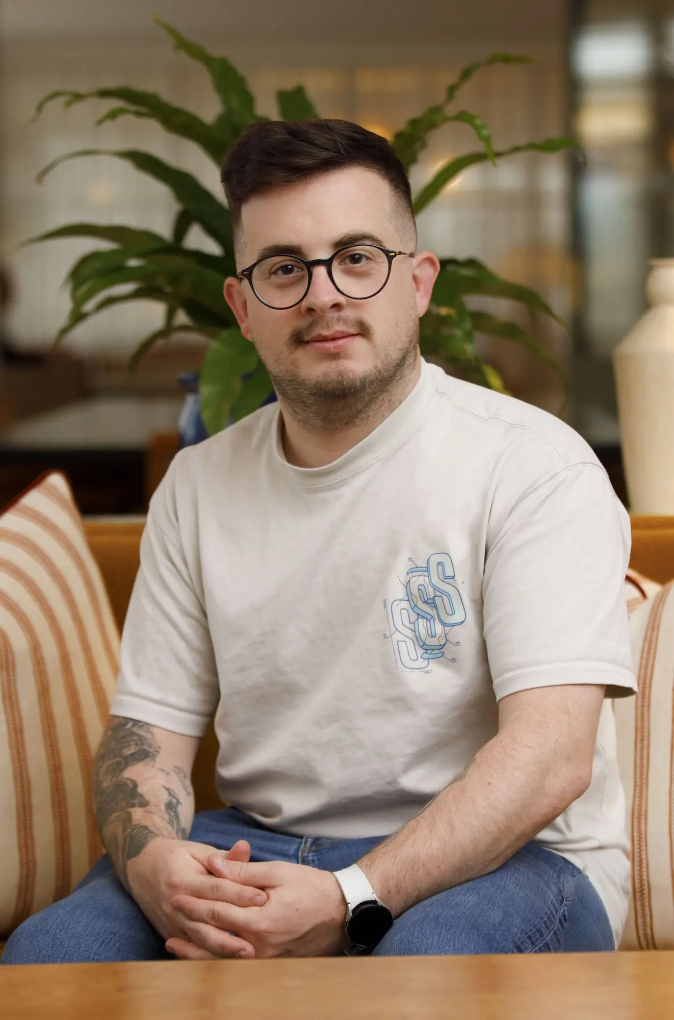 Man with short dark hair, glasses, and a tattoo on his left forearm sitting on a couch with striped cushions and a large green plant in the background, wearing a light-colored t-shirt and blue jeans.
