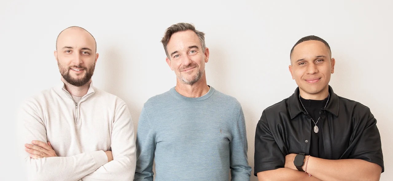 Three men standing against a white background; the man on the left wears a beige sweater with arms crossed, the middle man wears a light blue sweater and smiles slightly, and the man on the right wears a black shirt and jacket with arms crossed, also smiling.