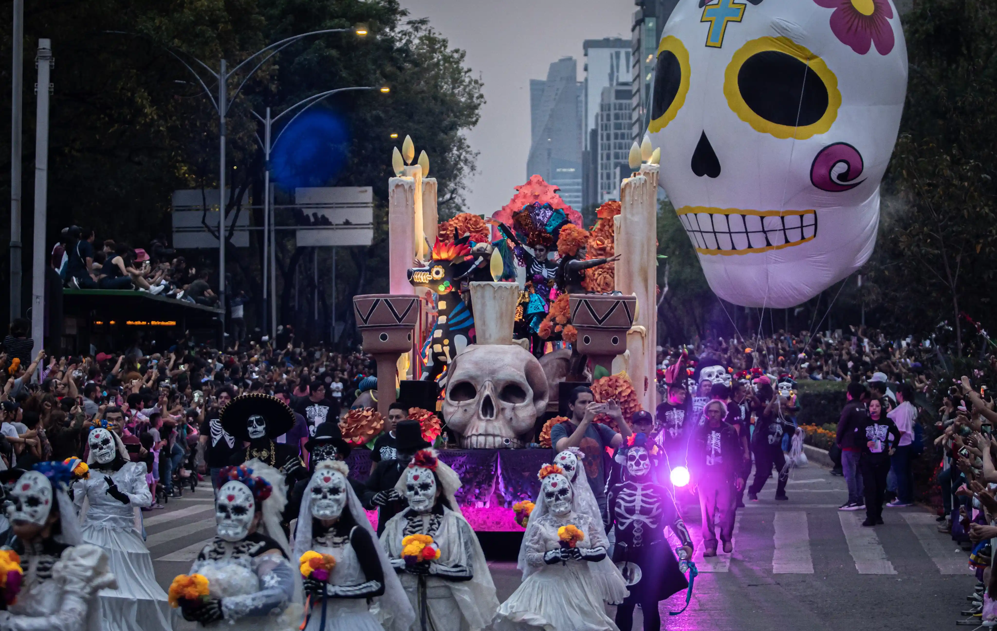 Day of the Dead parade with people in skeleton and Catrina costumes marching, accompanied by an elaborate float with a giant skull and a large skeleton balloon floating above, with skyscrapers in the background.