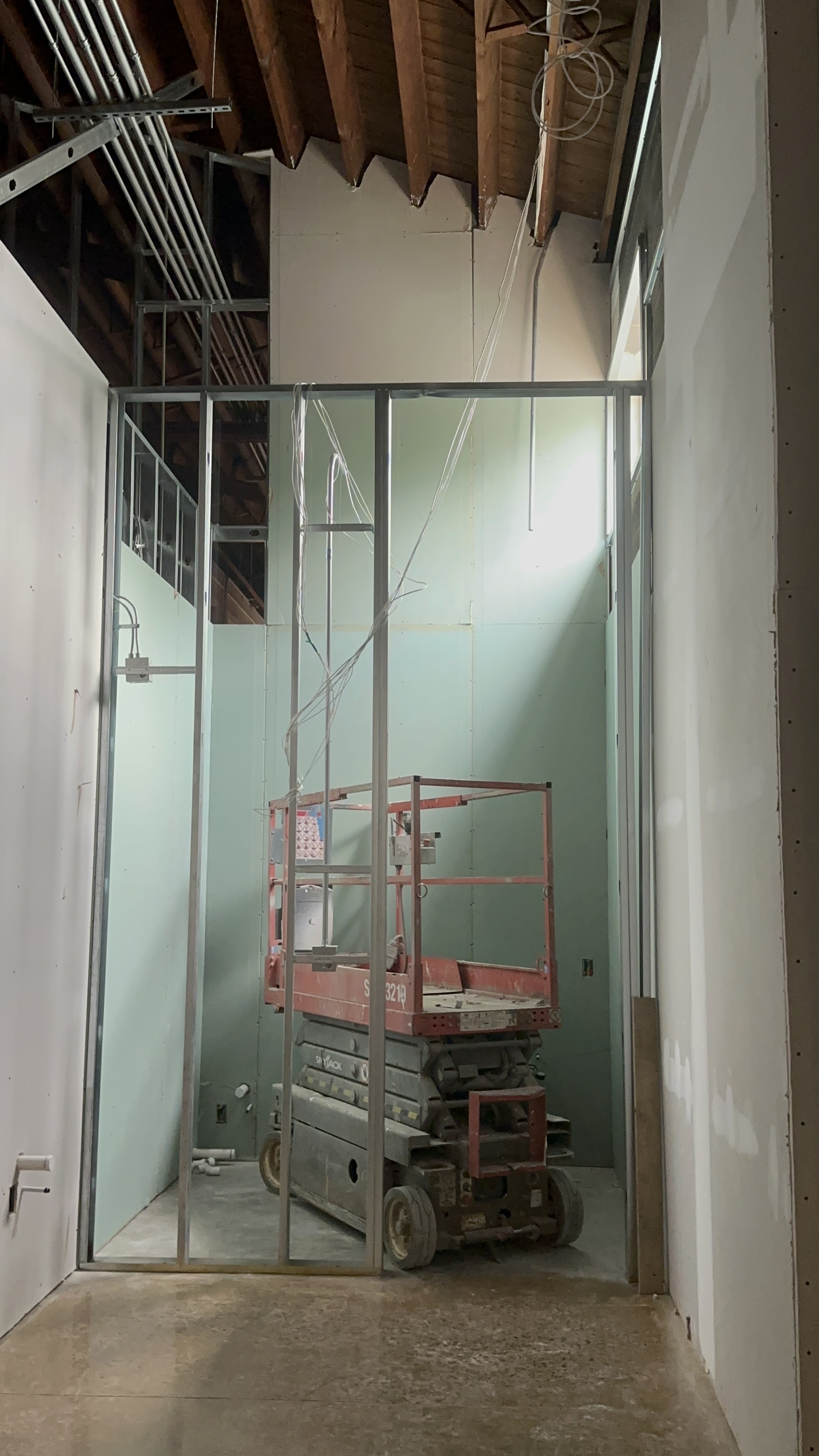 Red scissor lift inside a partially constructed room with exposed metal framing and green drywall panels.
