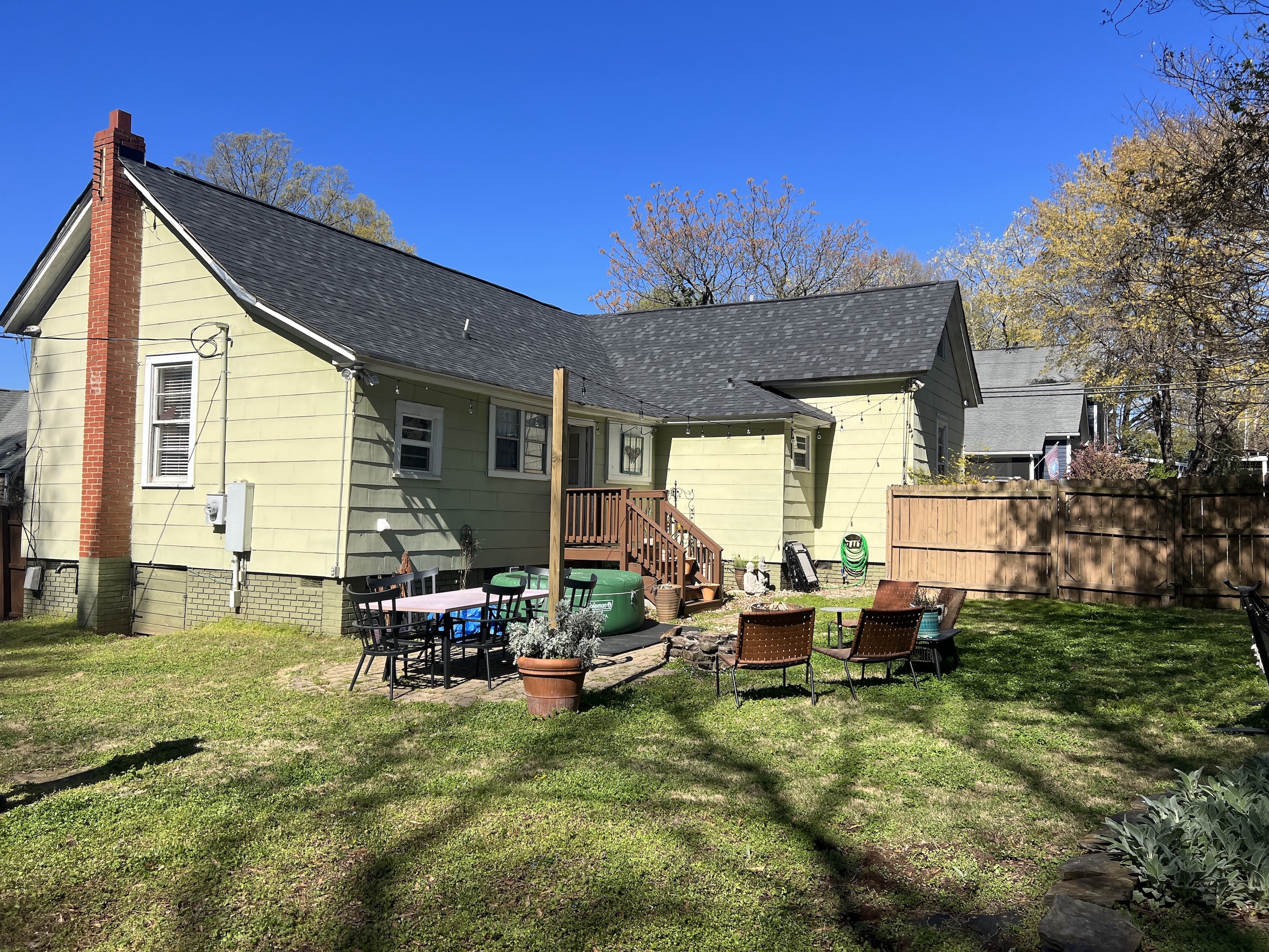 Backyard of a yellow house with a brick chimney, patio table with chairs, lawn chairs around a fire pit, and wooden fence under a clear blue sky.