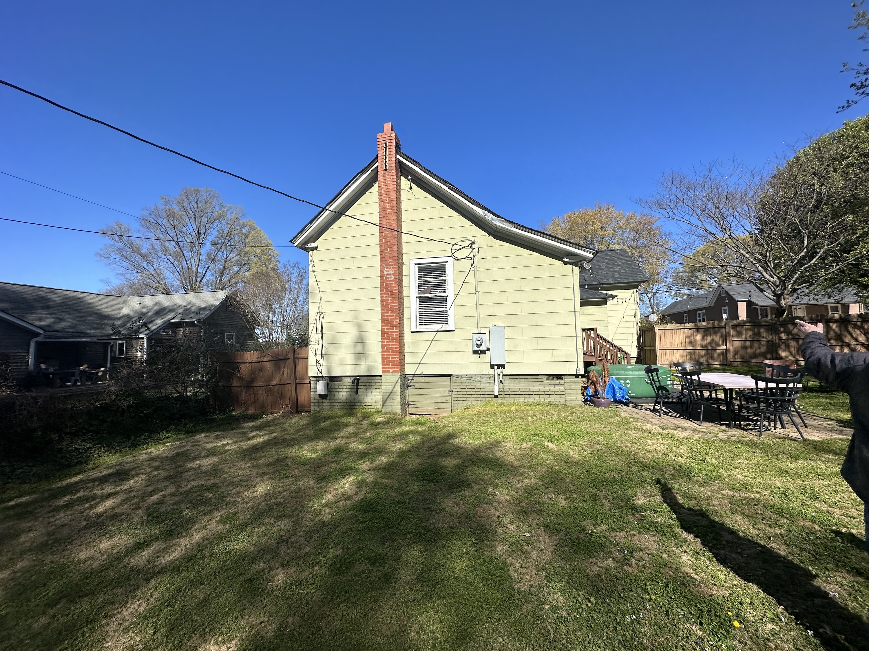 Side view of a yellow house with a red brick chimney, a window, and a wooden fence, with a patio set and grassy yard under a clear blue sky.