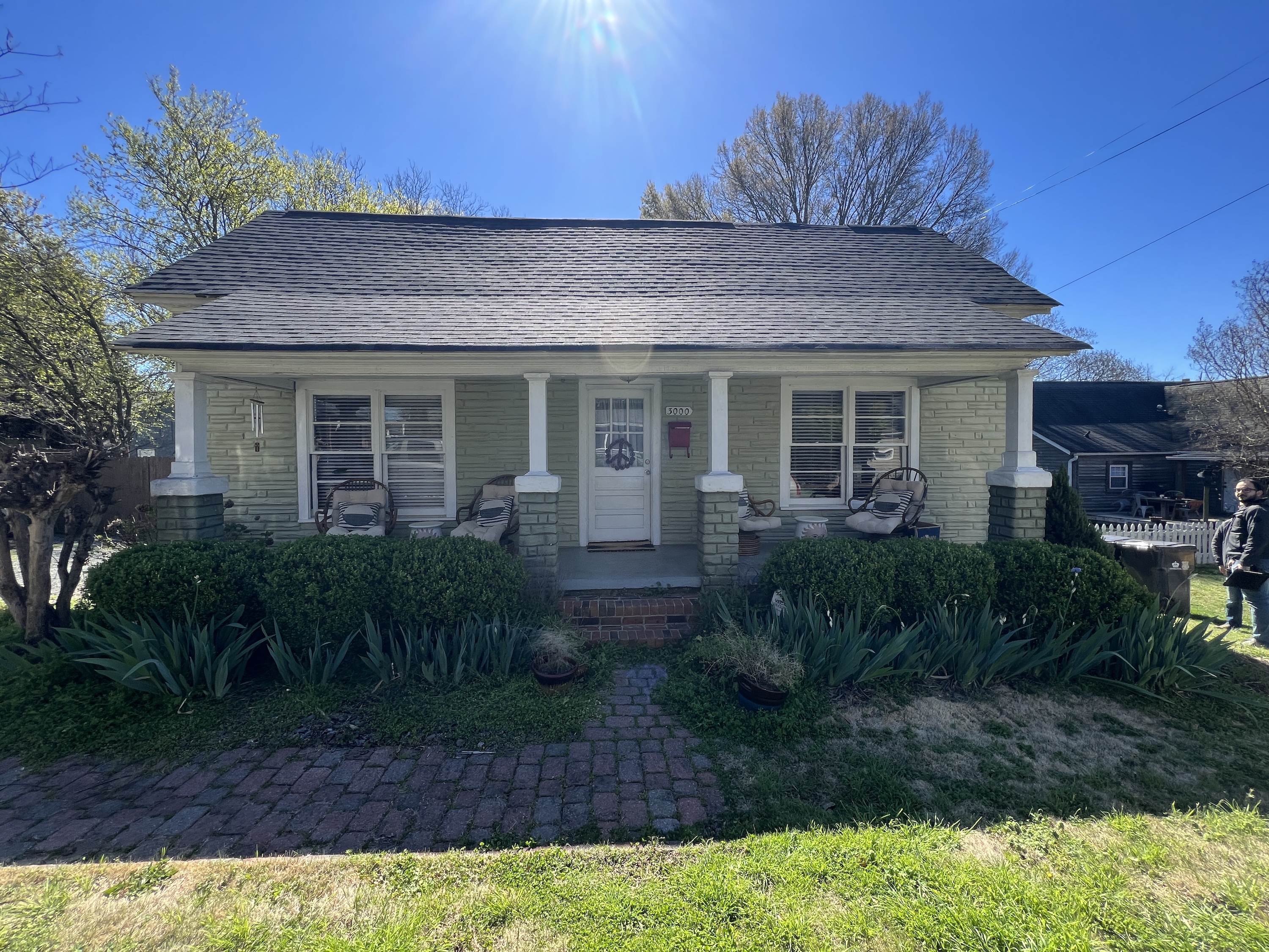 Single-story house with yellow siding, a front porch with four white columns, patio chairs, and bushes in front, under a clear blue sky.