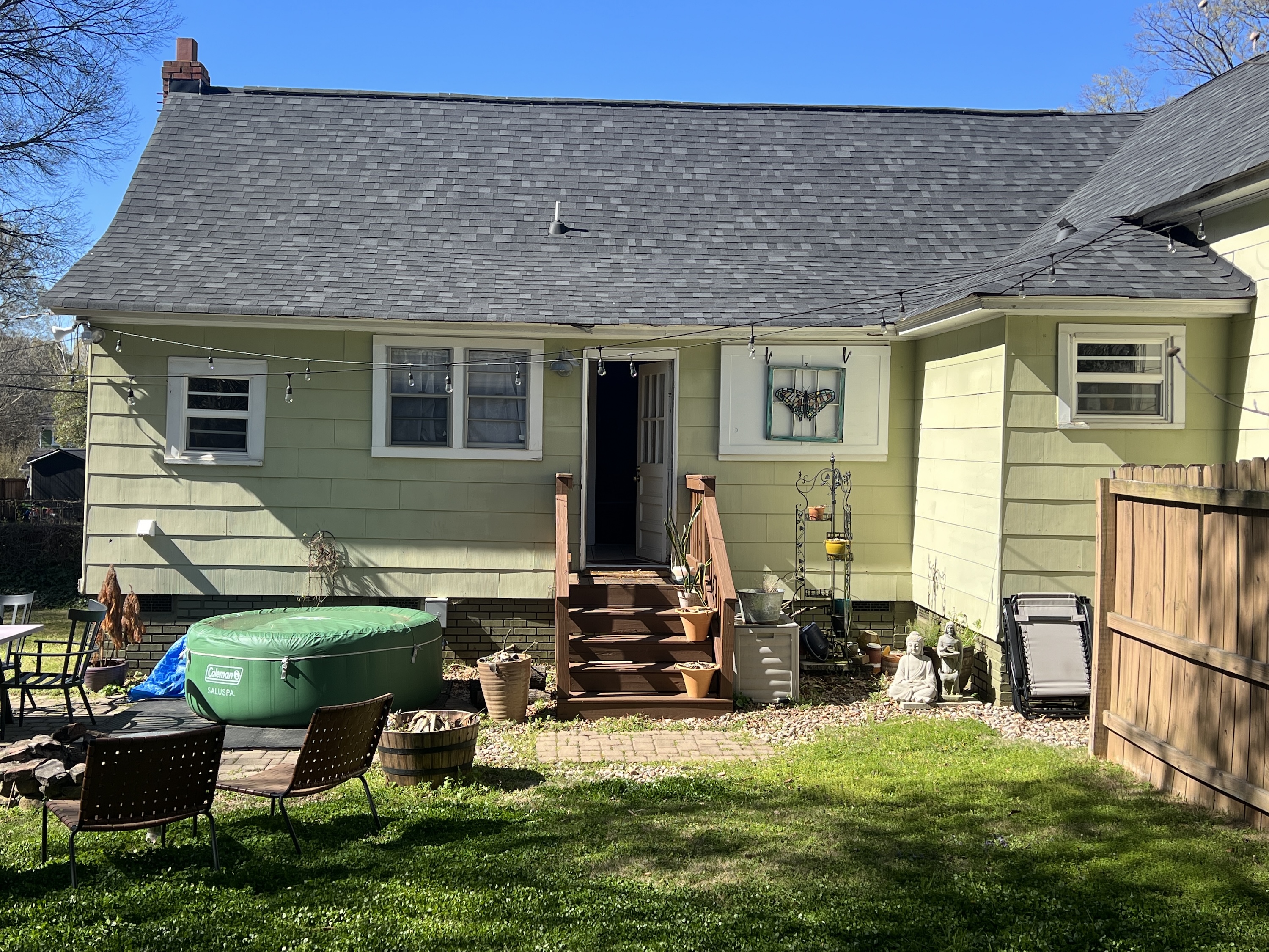 Backyard of a light yellow house with a wooden staircase, green inflatable hot tub, patio furniture, garden decorations, and a wooden fence.
