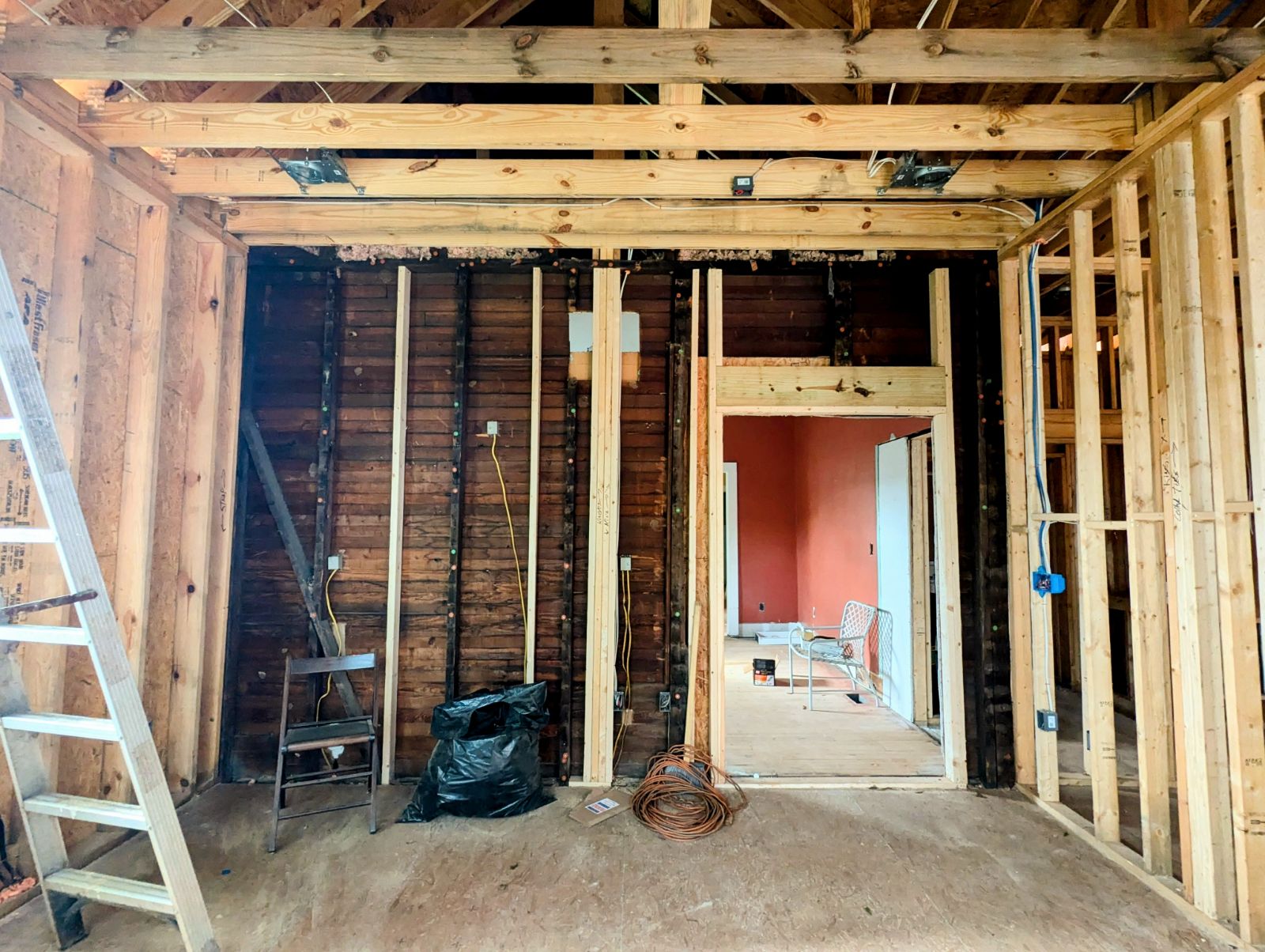 Interior view of an addition in progress. A wooden-framed room under construction with exposed beams, studs, a metal chair, a black trash bag, coiled cable, and a ladder on the left.
