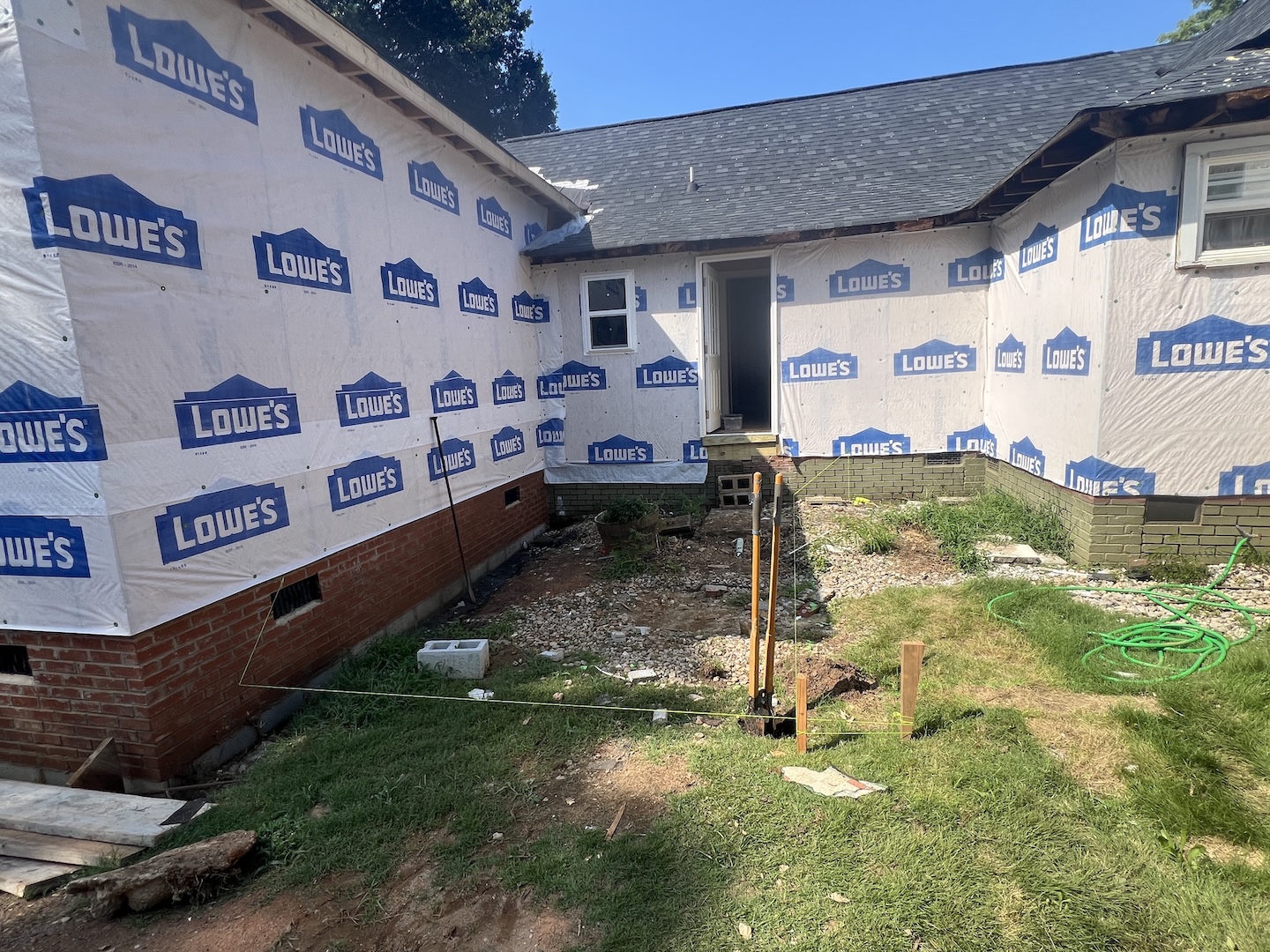 House exterior under renovation with Lowe's branded house wrap, an open door, construction tools in the yard and a green garden hose on the grass.