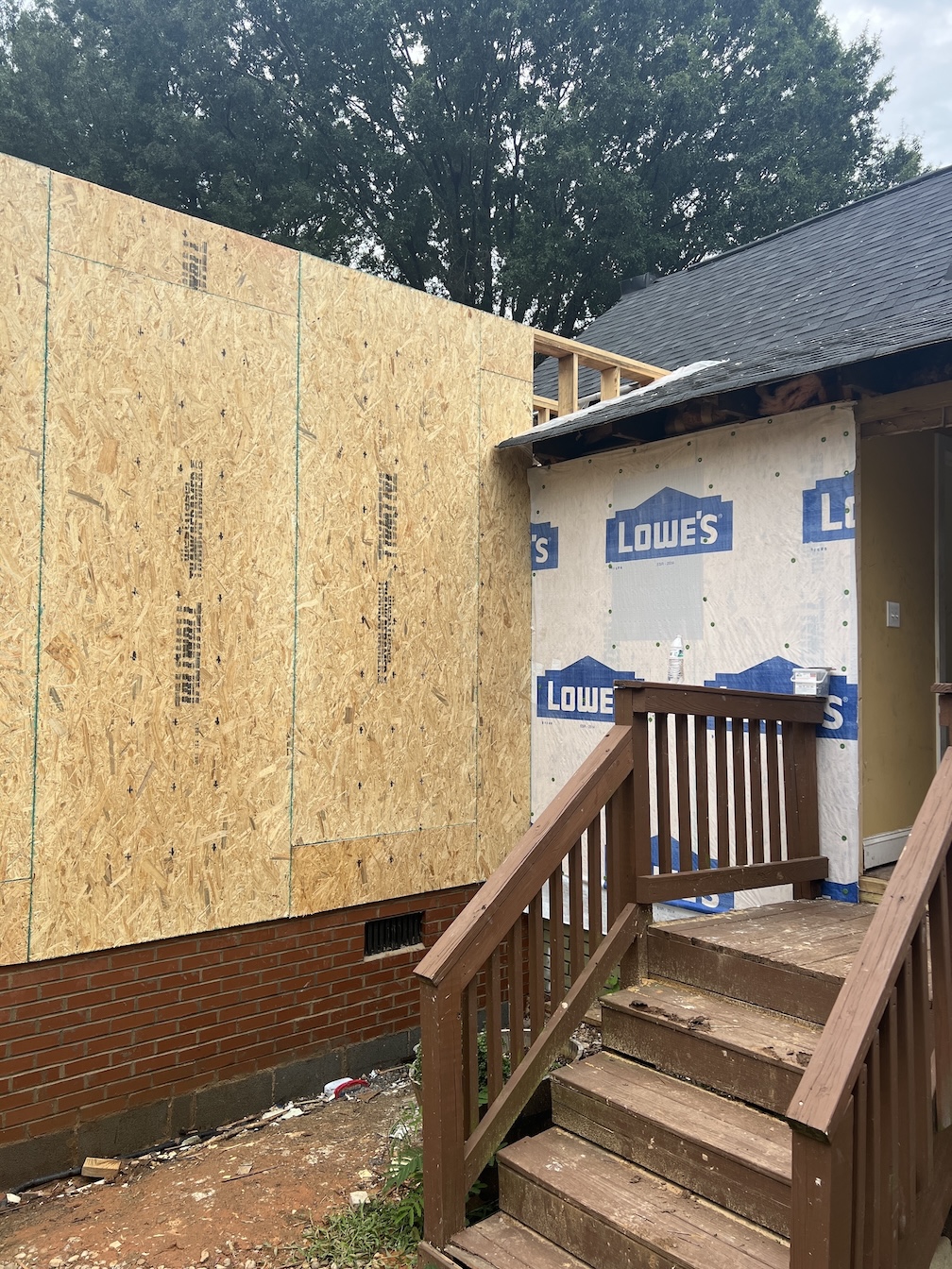 Wooden stairs with railing leading to a house entrance under renovation, with new plywood and house wrap on the exterior wall.