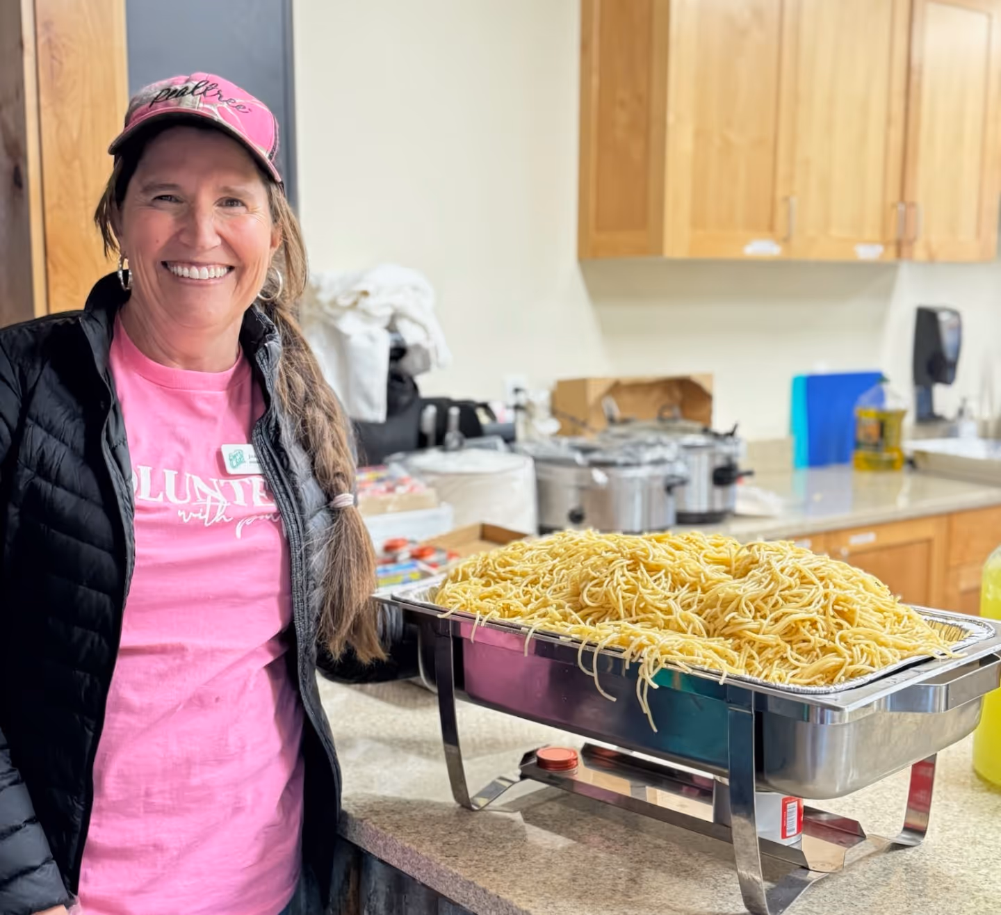 A woman in a pink hat and shirt smiles while standing behind a large catering pan filled with pasta in a commercial kitchen.