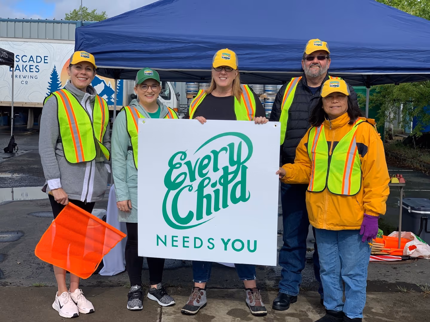 Three volunteers in safety vests and hard hats hold up a large green and white "Open House" sign.