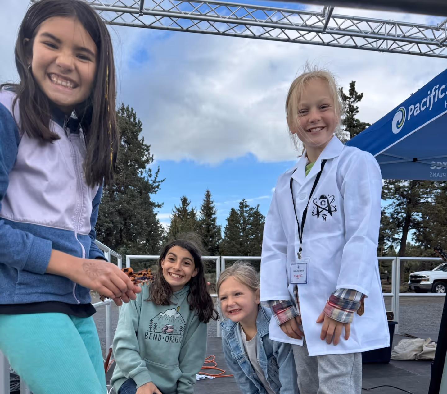 A smiling woman in a white lab coat poses with three young children in front of an event tent.