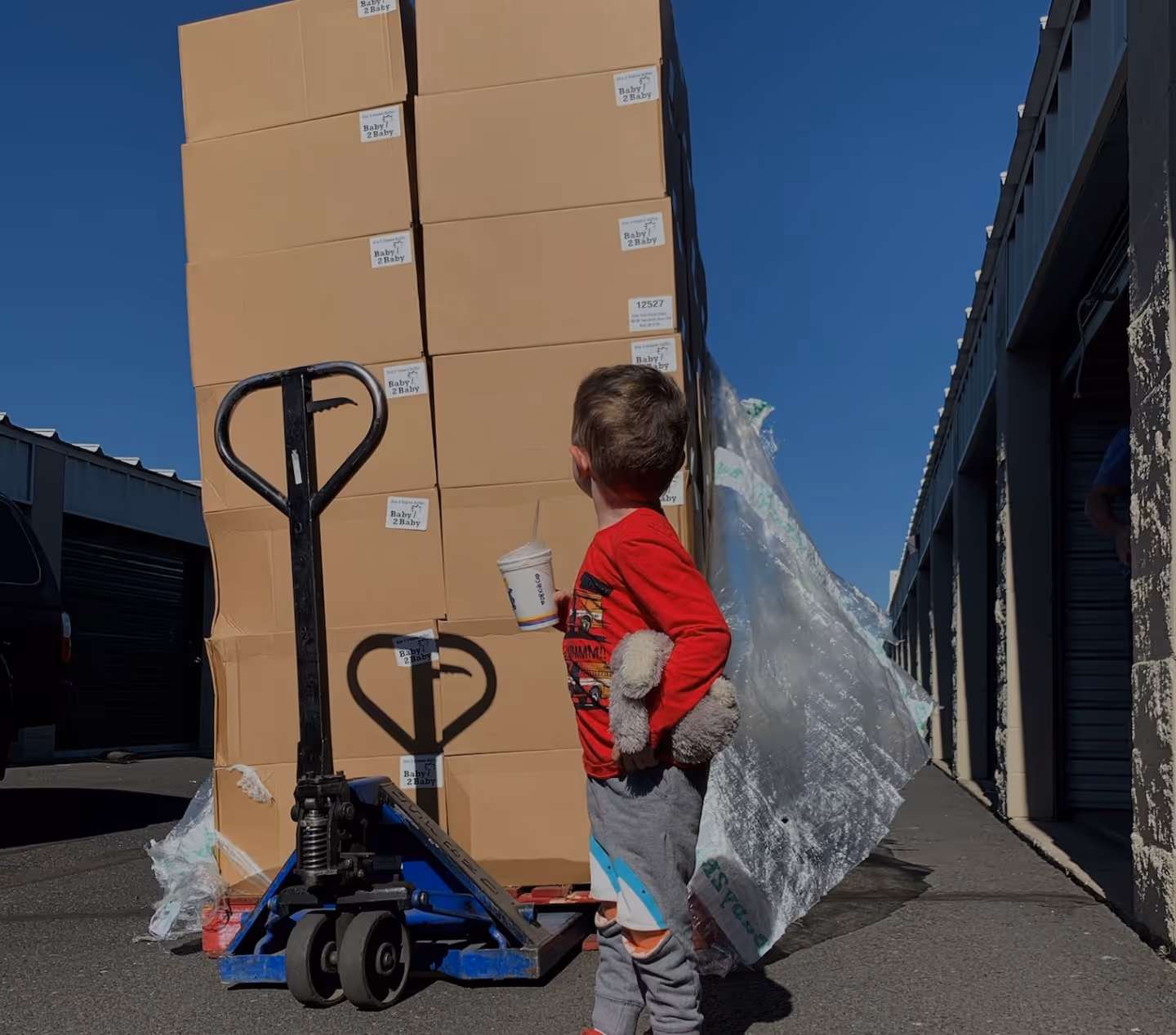 A small child in a red shirt stands next to a hand truck loaded with boxes outdoors.