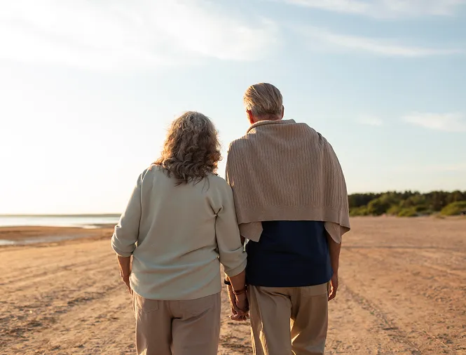 An elderly couple holding hands and walking on a sandy beach during sunset.