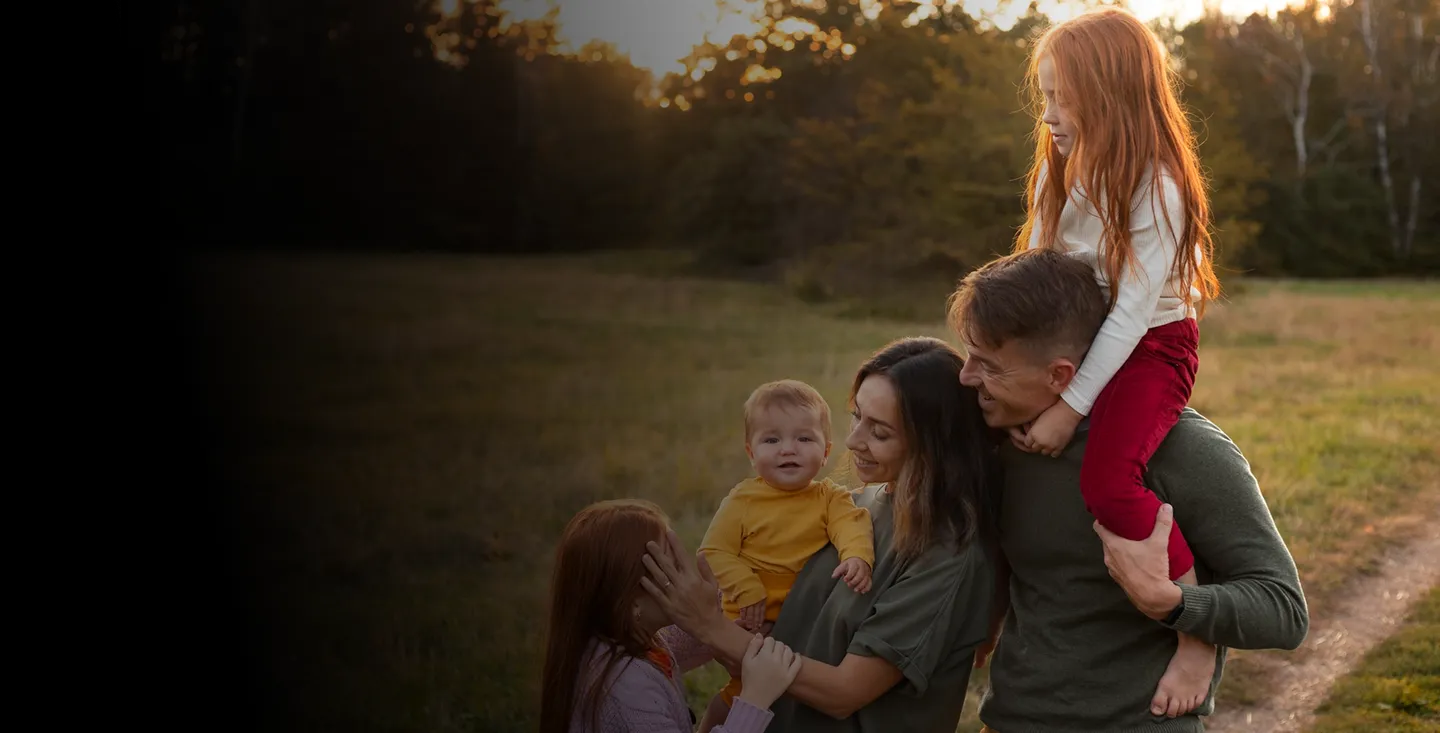 Happy family outdoors at sunset with father carrying daughter on shoulders, mother holding baby, and another child touching baby’s face.